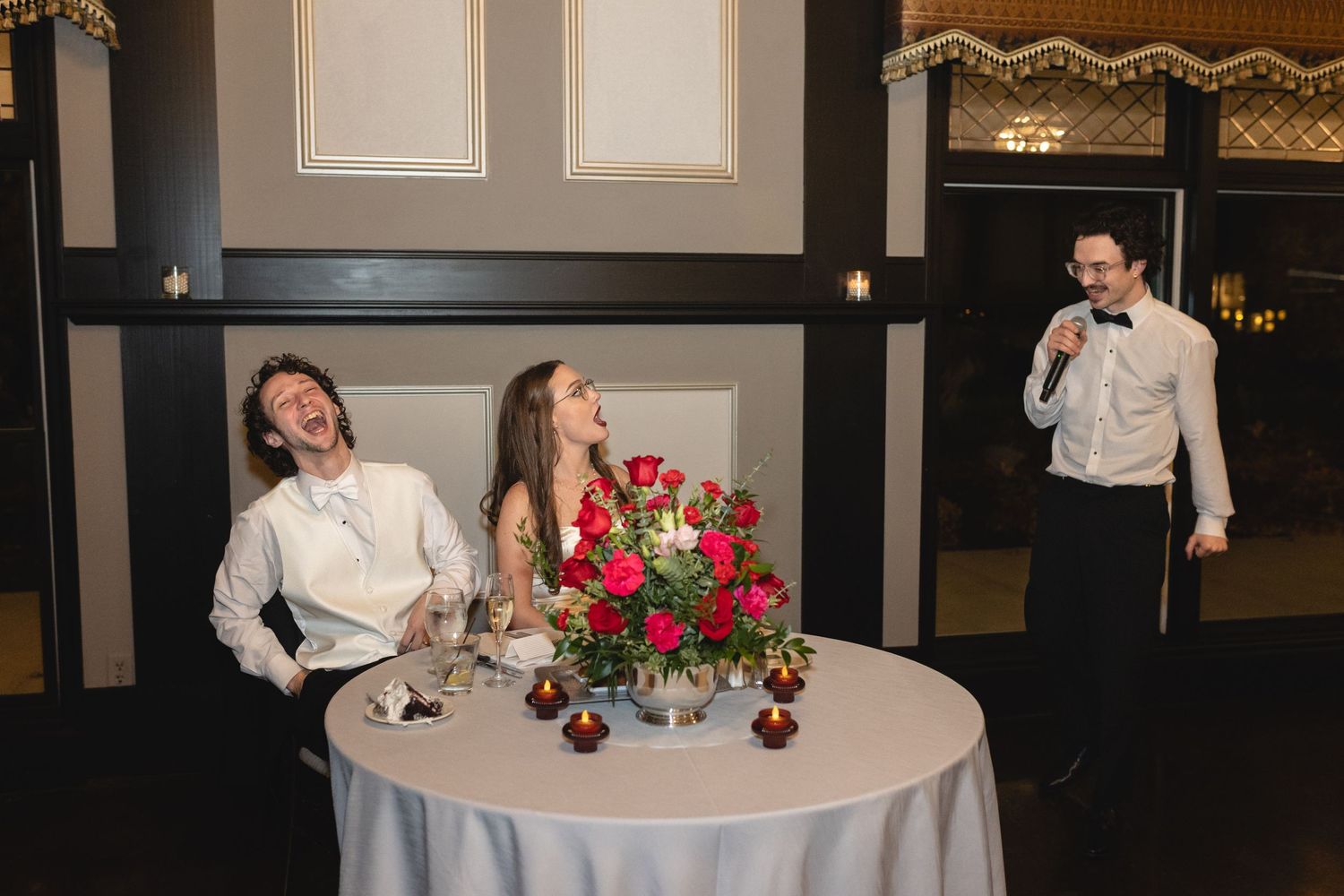 Candid moment at an elegant reception with laughter around a table decorated with vibrant red floral centerpiece.