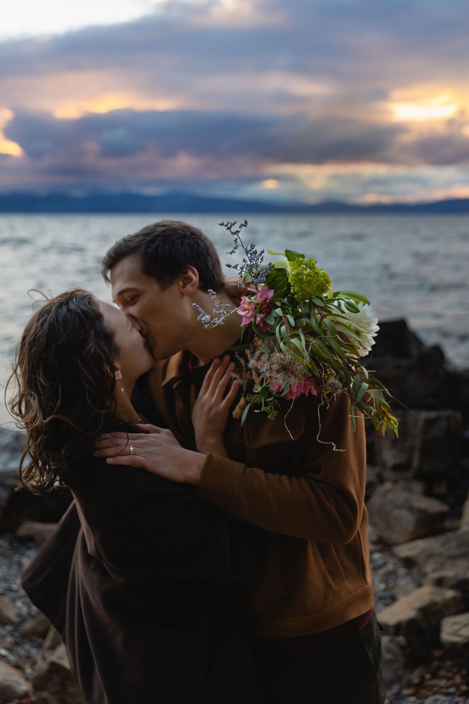 A tender moment shared on a rocky beach at sunset with a beautiful bouquet of flowers and dramatic cloudy sky.