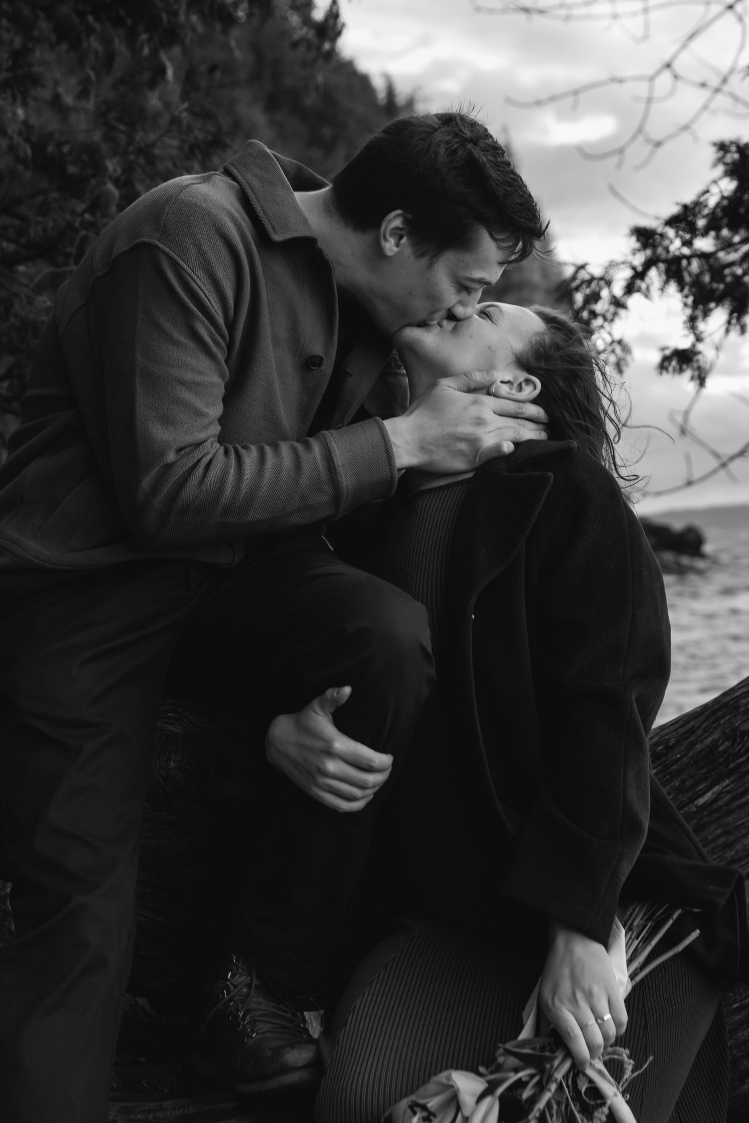 Intimate black and white photo of a couple sharing a romantic moment by the water at sunset.