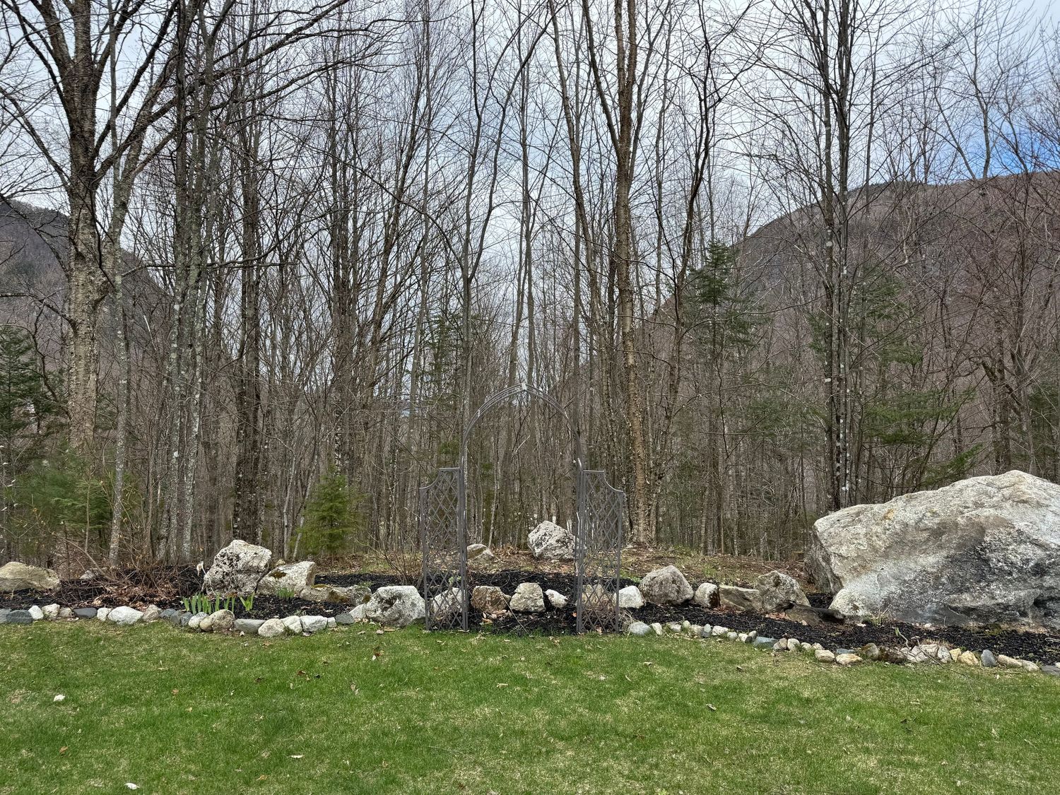 A rustic stone wall stretches along the edge of a wooded area with bare trees and green grass in early spring.