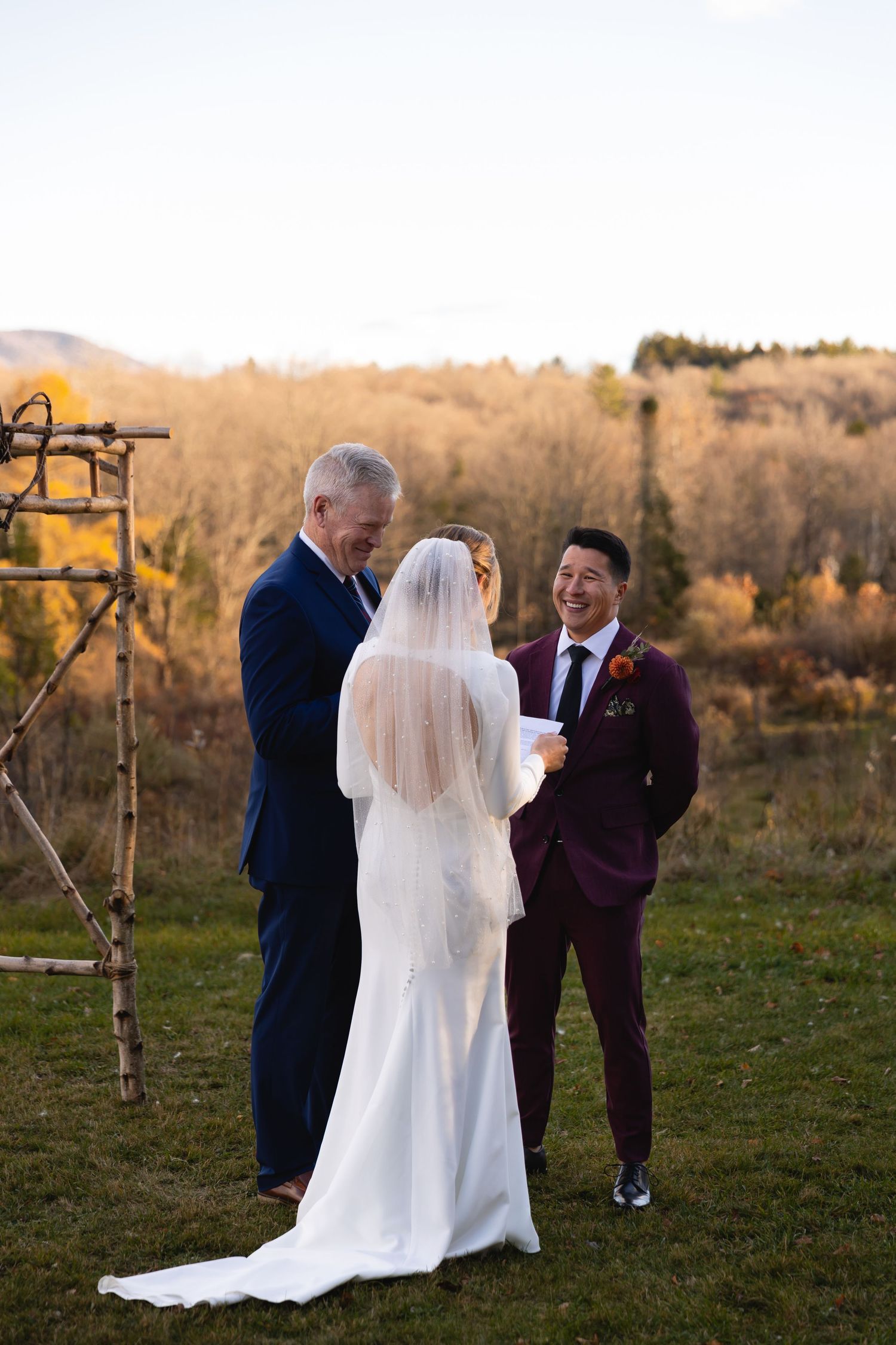 An outdoor wedding ceremony in autumn with a couple and officiant standing under a rustic wooden arch during golden hour.