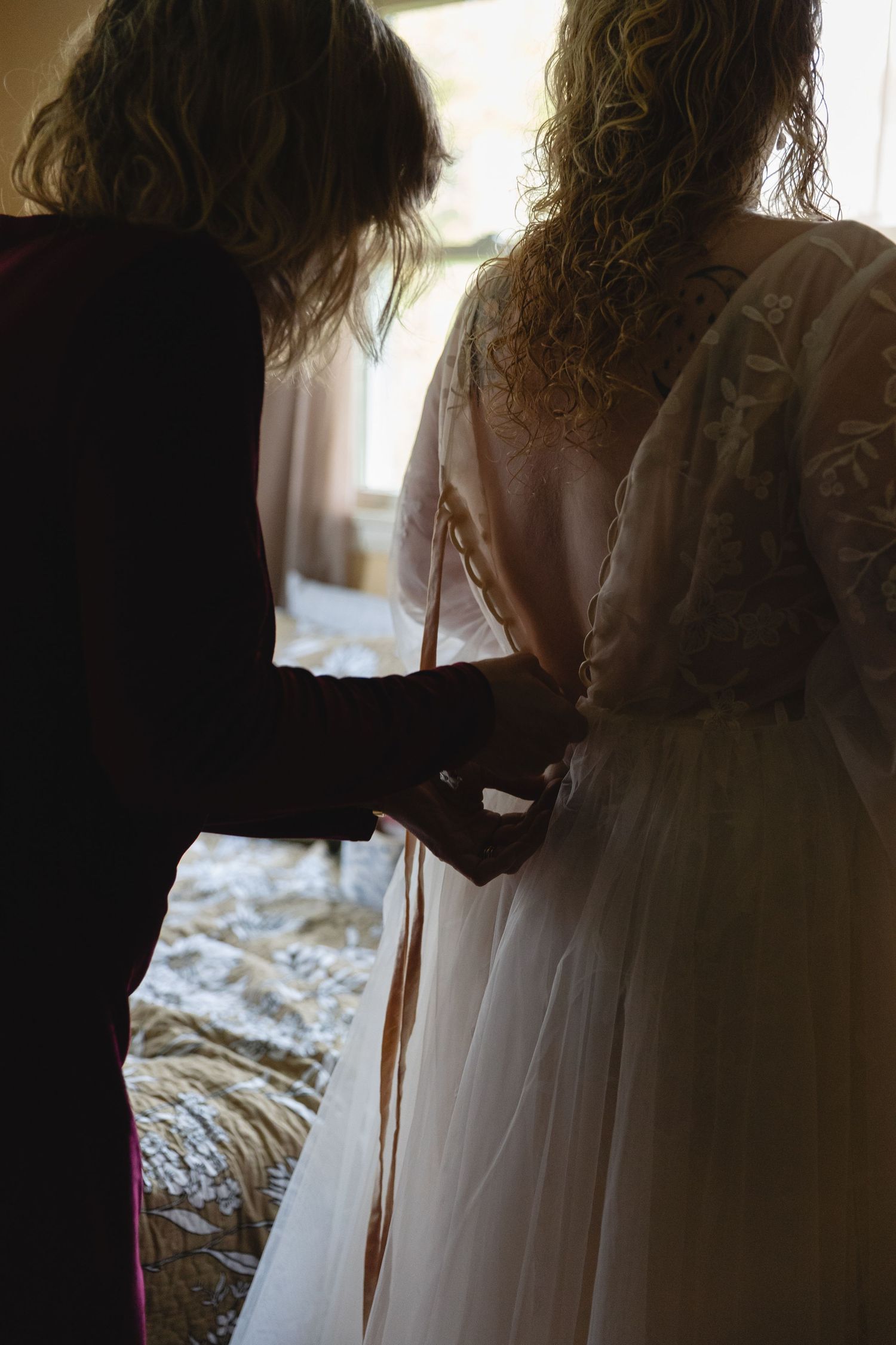 Silhouetted figure helping to button up a lace wedding dress in soft window light.