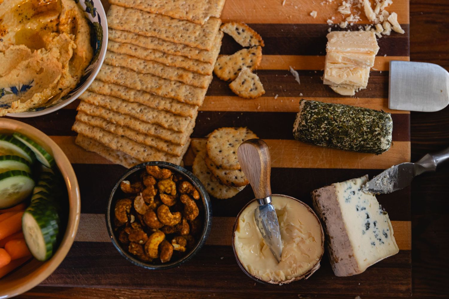 Overhead view of a charcuterie board with crackers, blue cheese, nuts and fresh vegetables on a wooden surface.