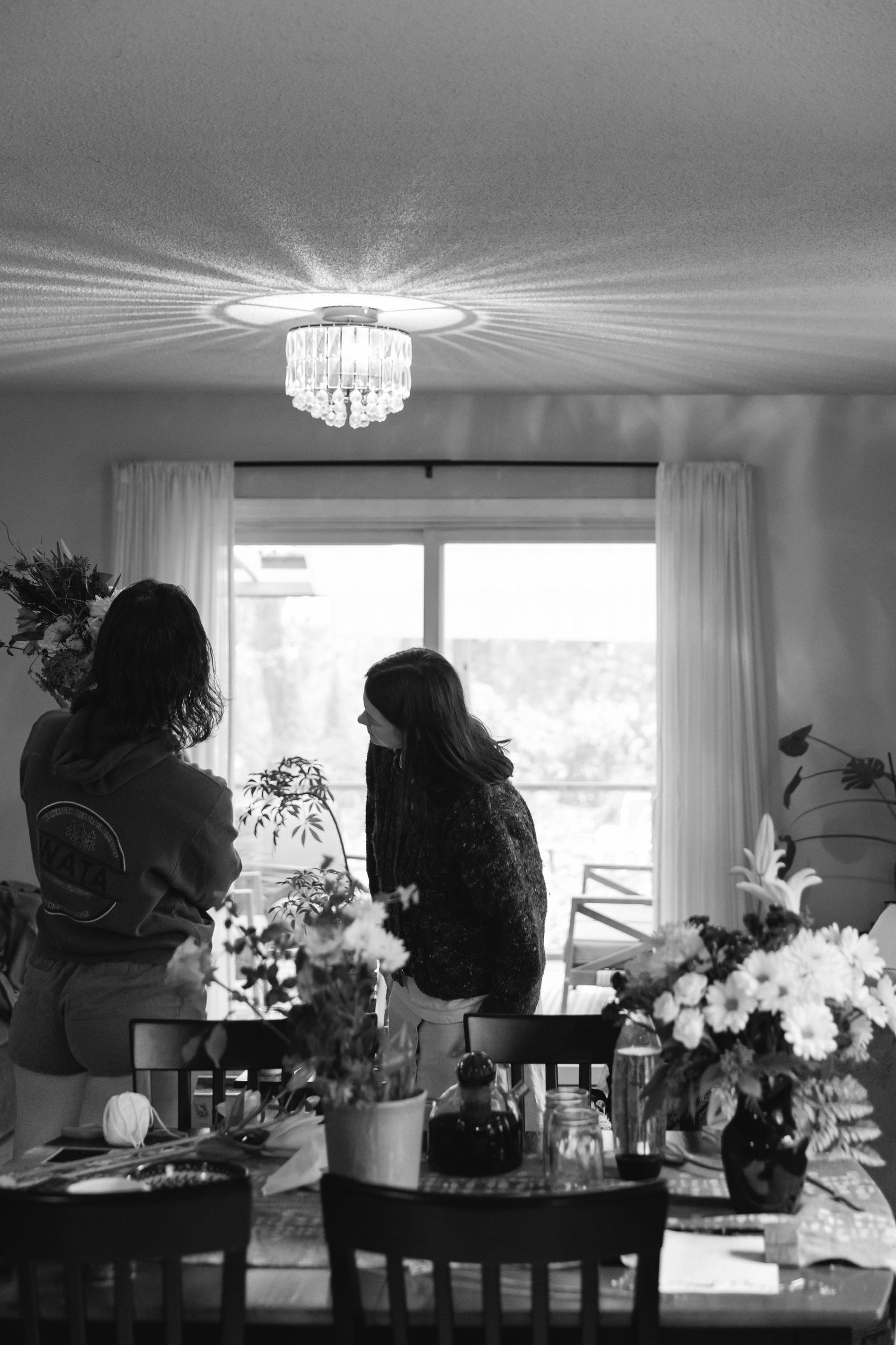 Black and white photo of a dining room with crystal chandelier, floral arrangements, and two silhouettes by a window.