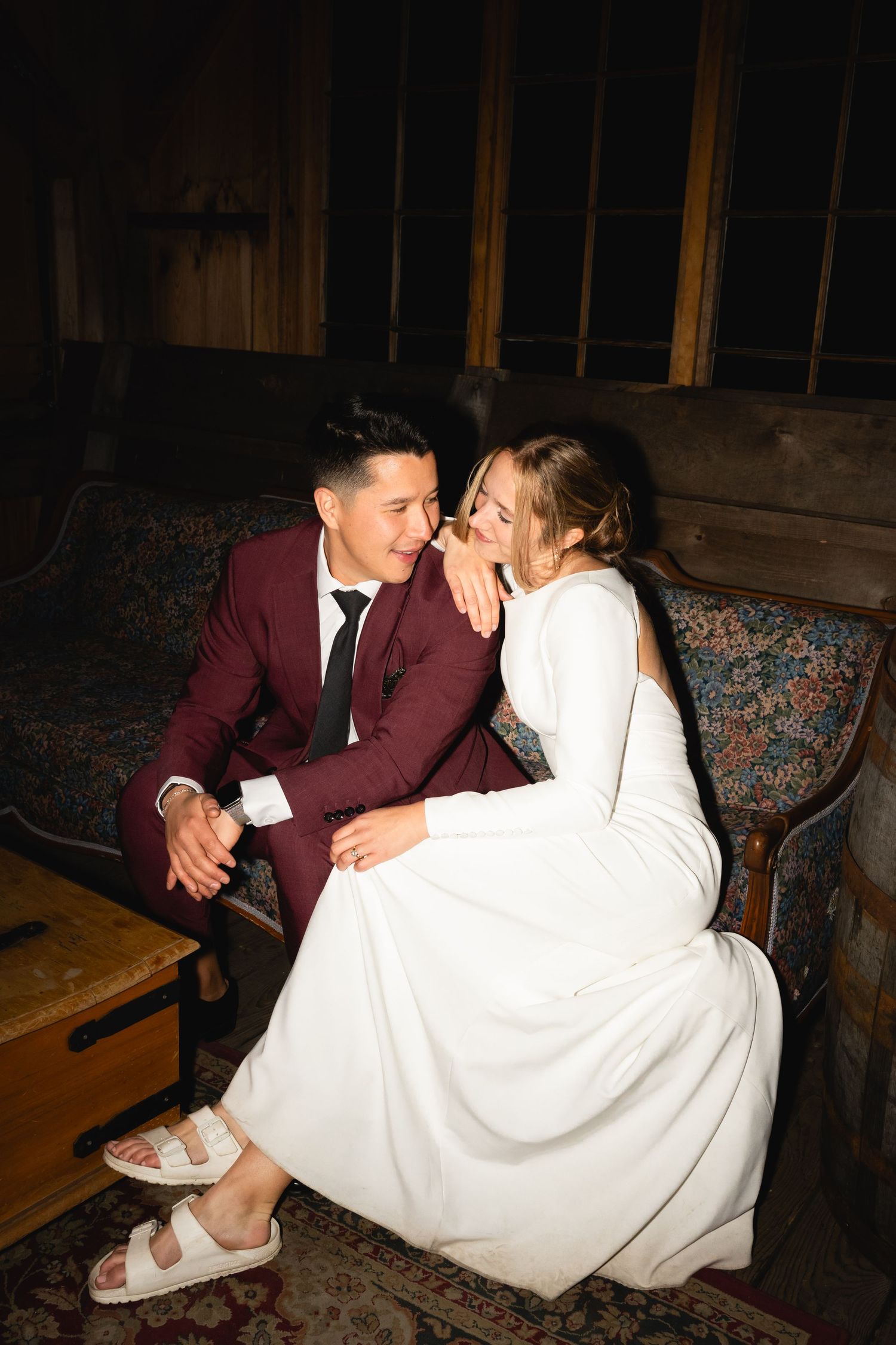 A couple shares an intimate moment on a vintage couch, with the bride wearing sandals and a long white dress.