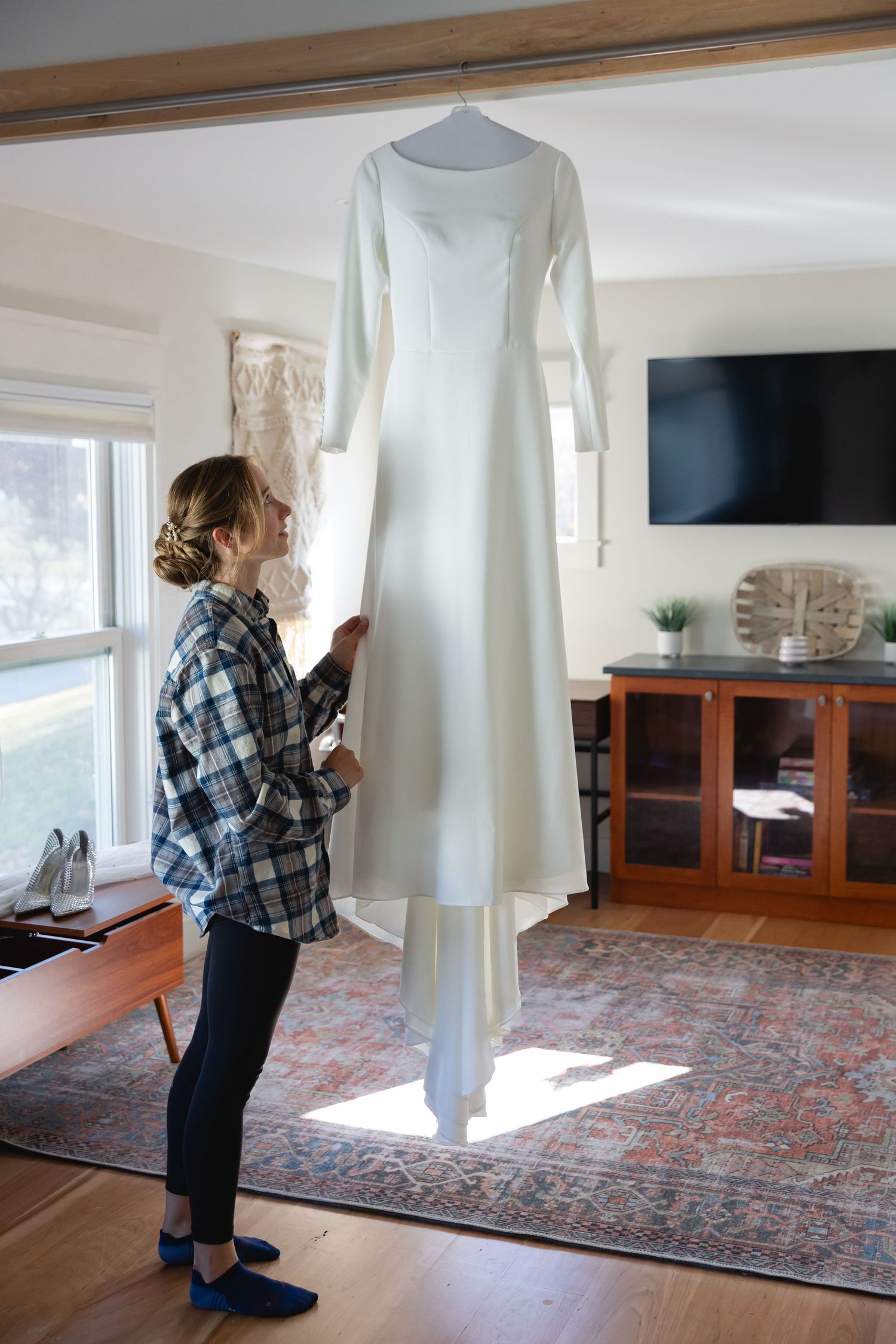 A white wedding dress with long sleeves hangs in a sunlit room with wood floors and an oriental rug.