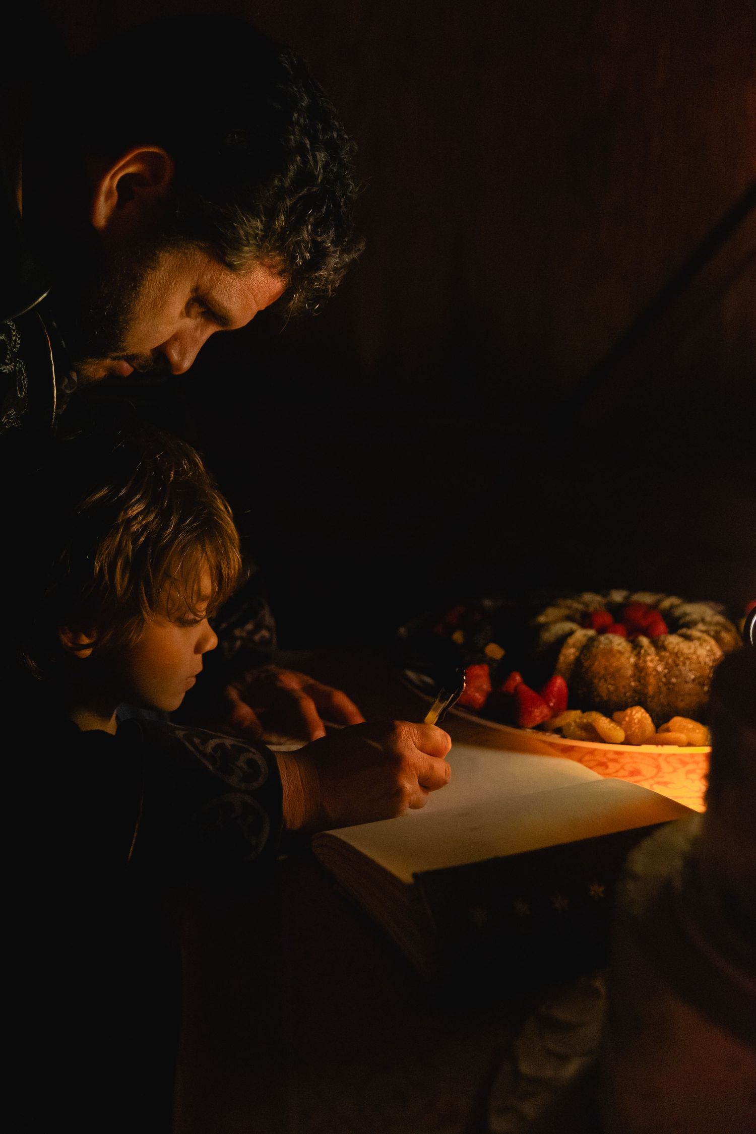Father and child writing together by candlelight beside a festive cake in a dark room.