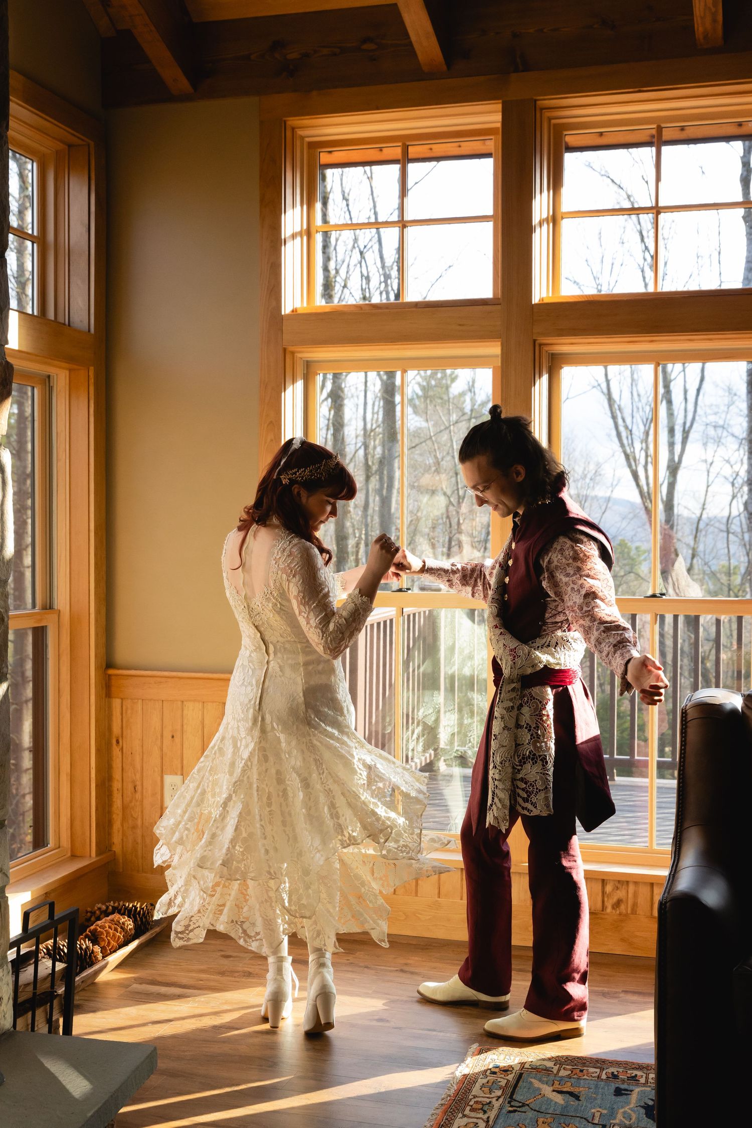 Couple in unique wedding attire dancing by large sunny windows with mountain views in background.