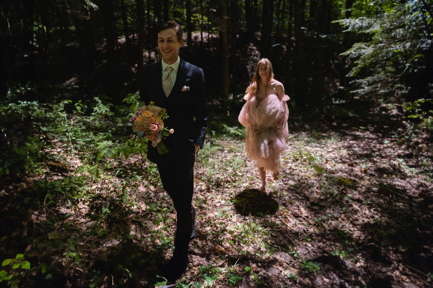 Groom walks through sunlit forest holding bouquet as bride follows in a pink dress behind.