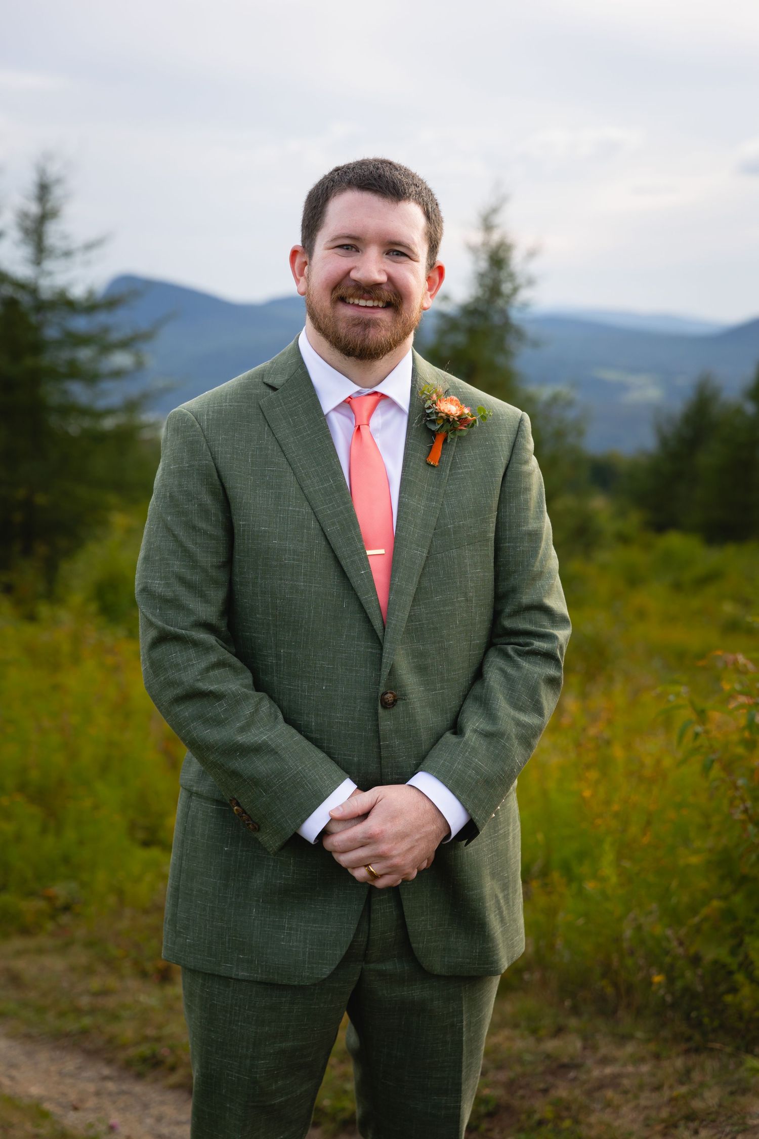 A person wearing a grey suit and coral tie stands outdoors against a mountain landscape background.