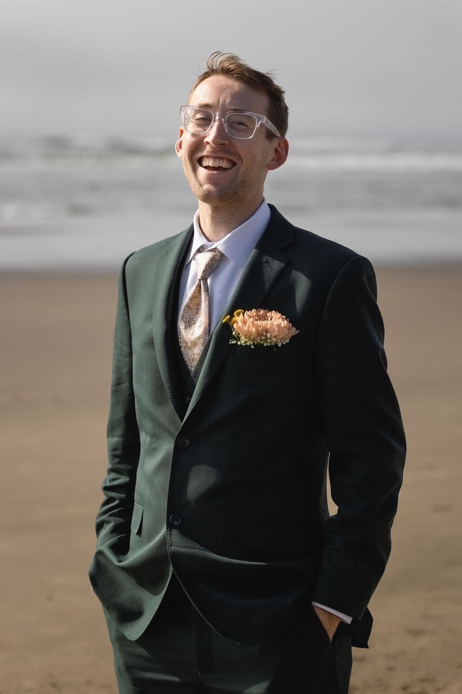 A joyful groom in a forest green suit and glasses smiles on a beach with a peach boutonniere.