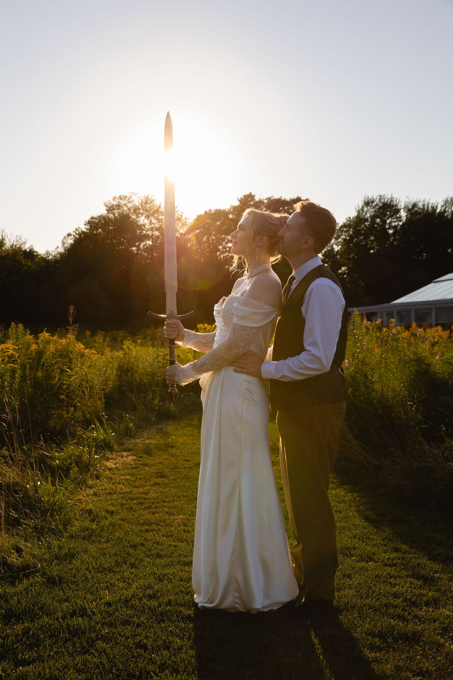 A couple in wedding attire stands together in a golden sunlit field during sunset, creating silhouettes against the sky.
