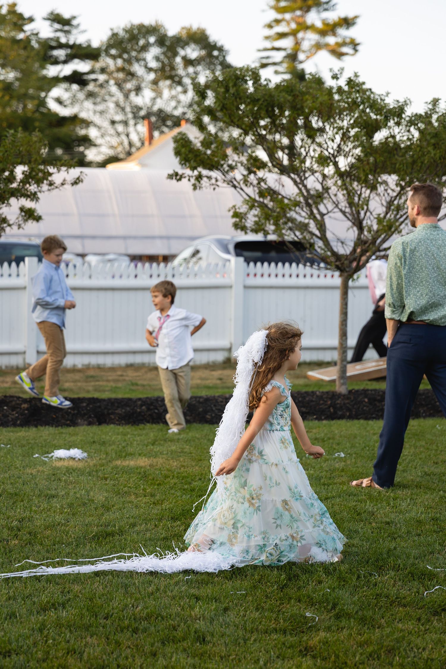 Children play and run on a grassy lawn near a white picket fence at sunset while wearing formal attire.