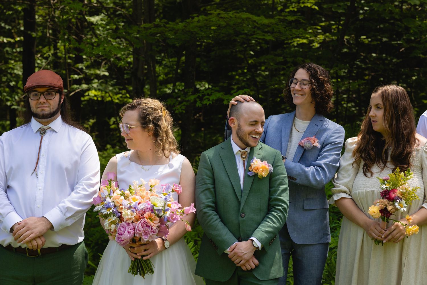 A wedding party stands in a line outdoors surrounded by lush green trees and forest scenery.