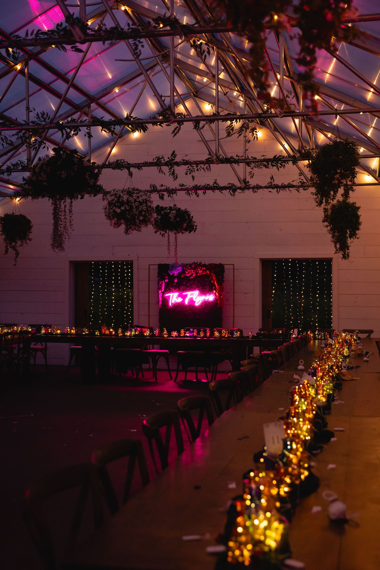 Indoor wedding reception venue decorated with hanging lights, greenery and pink neon sign creating a romantic ambiance.
