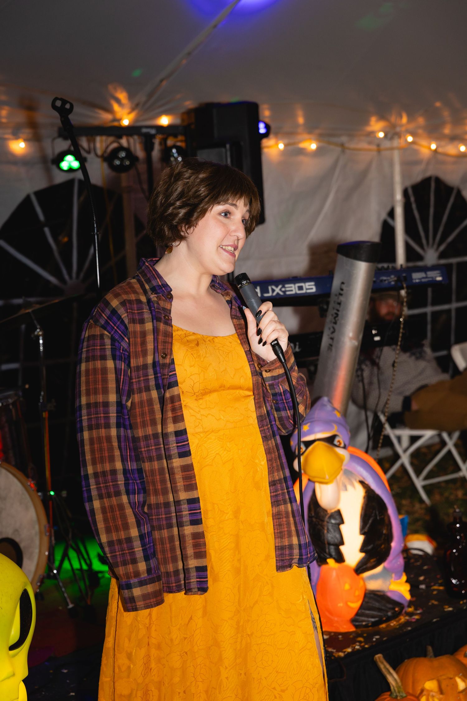 A performer in a yellow shirt and plaid flannel sings into a microphone at a dimly lit indoor venue.