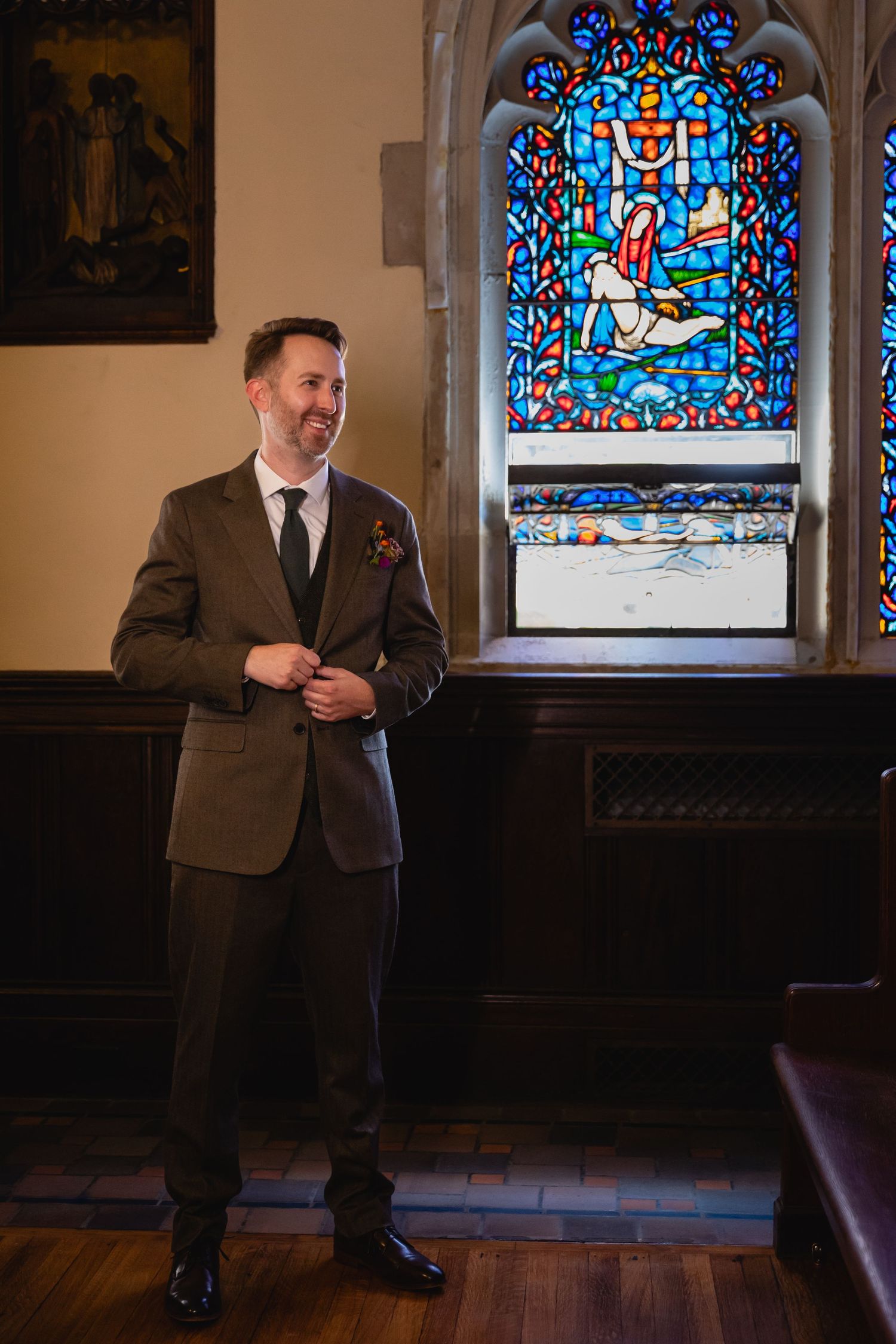 A person in a brown suit sits on a wooden church pew beneath colorful stained glass windows.