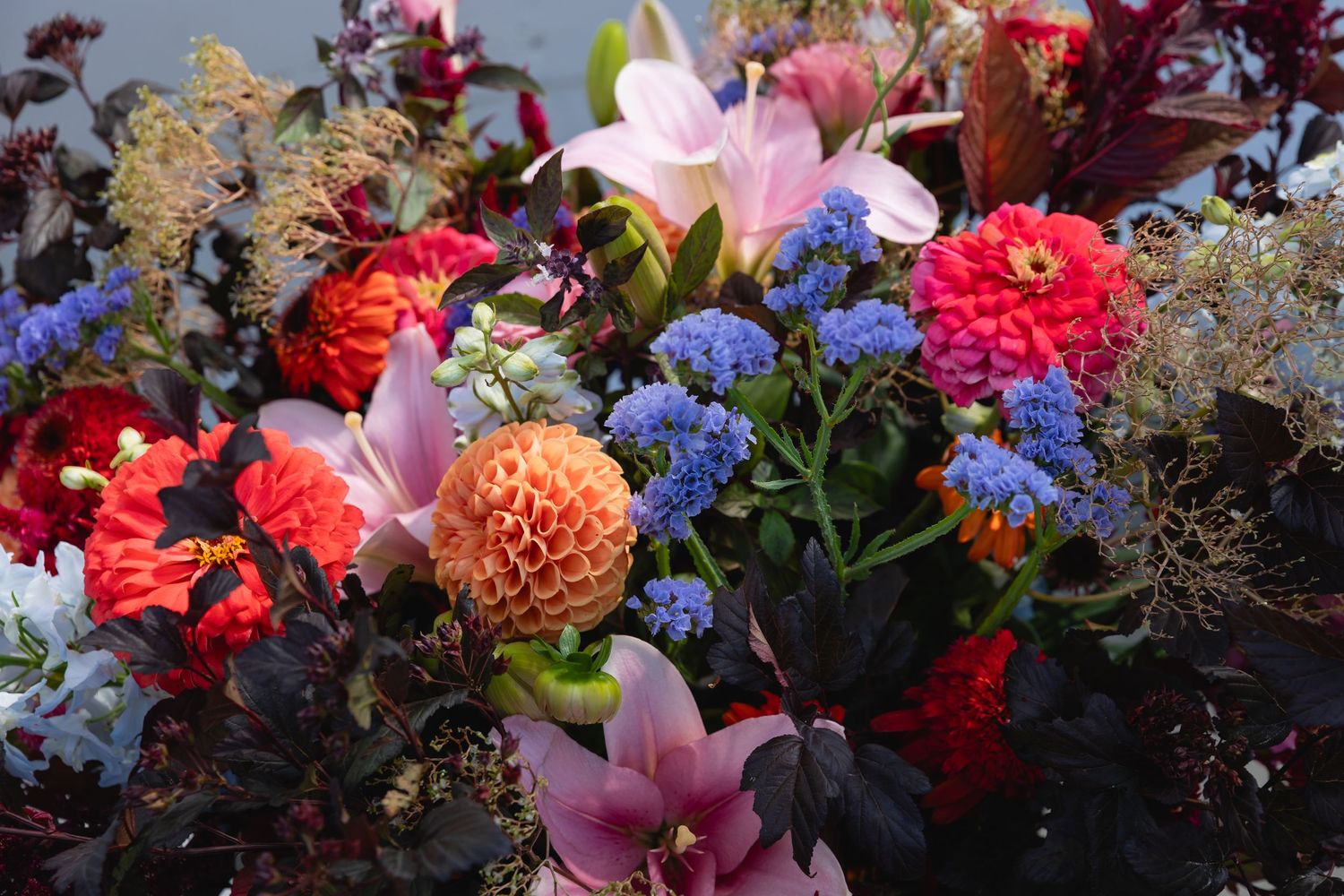 A vibrant floral arrangement featuring pink lilies, red roses, blue delphiniums, and peach dahlias in a rustic setting.