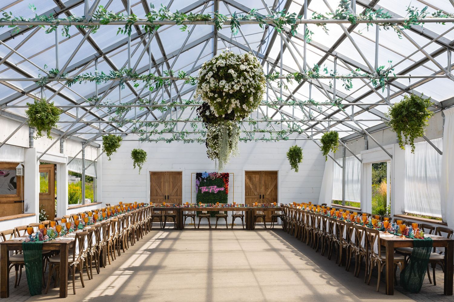 A panoramic view of a rustic greenhouse wedding venue with wooden tables and hanging greenery decorations.