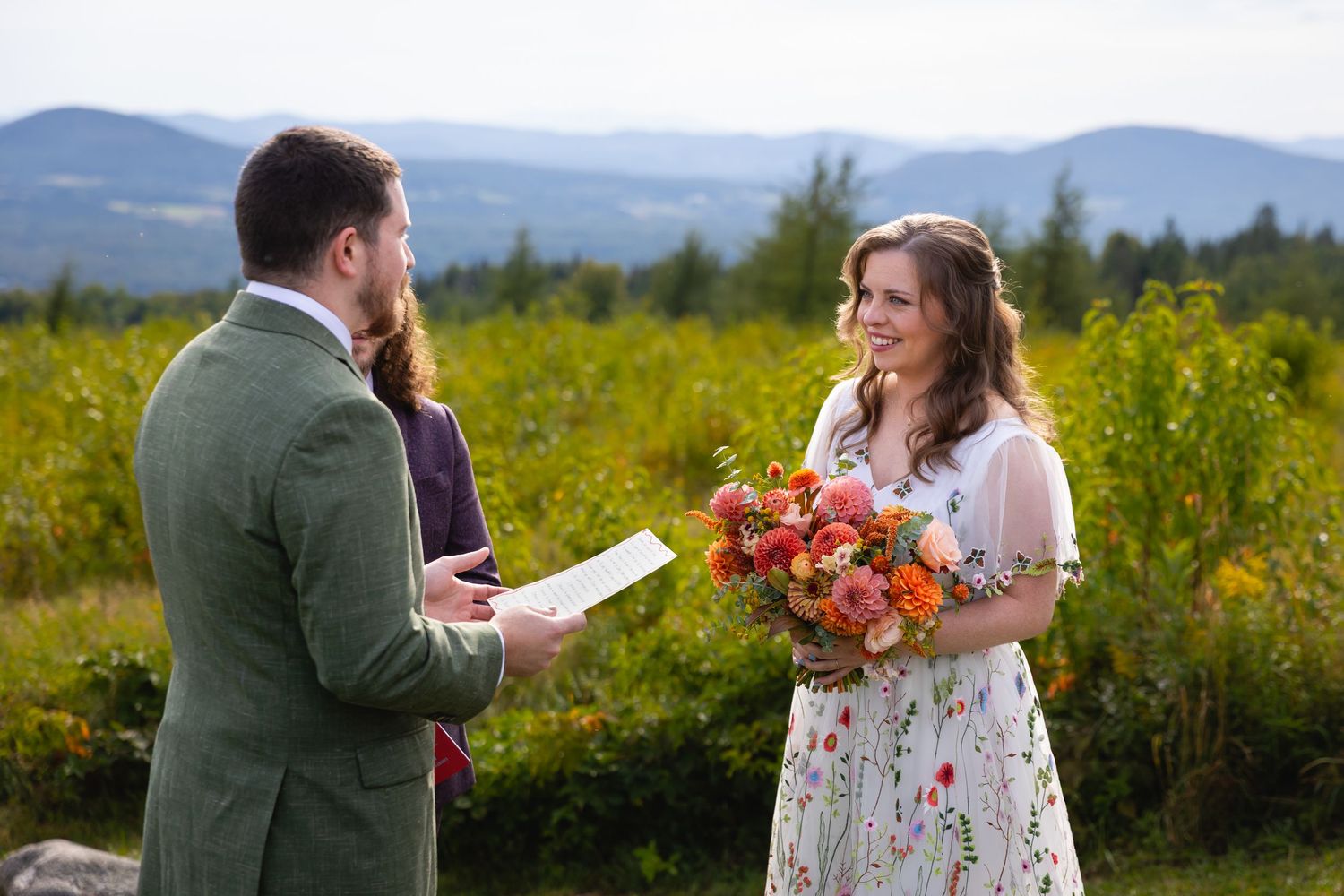 A couple stands in a sunny meadow with mountains in the background during a wedding ceremony.