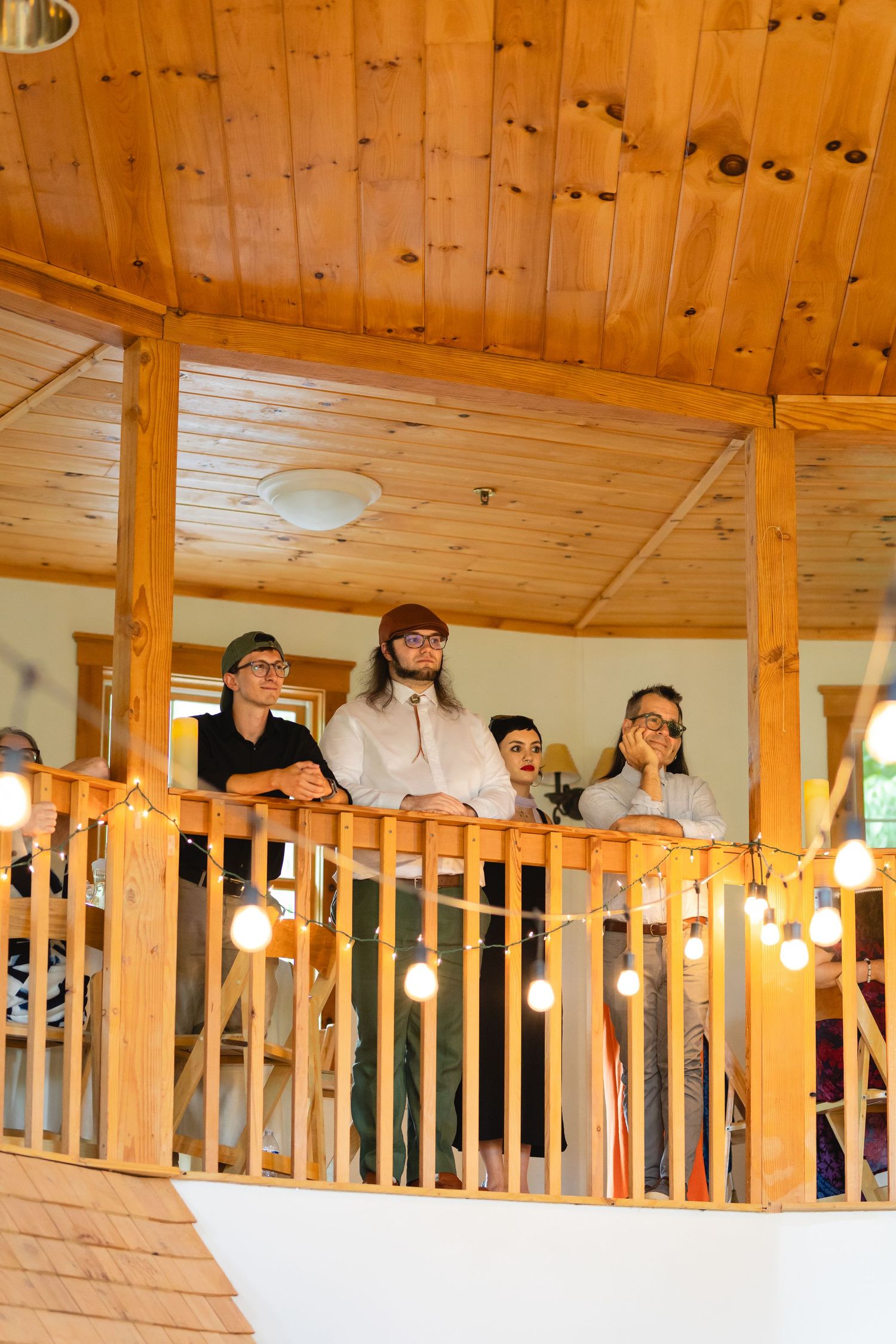 Wedding guests peer over a rustic wooden balcony railing in a lodge-style venue with warm lighting.