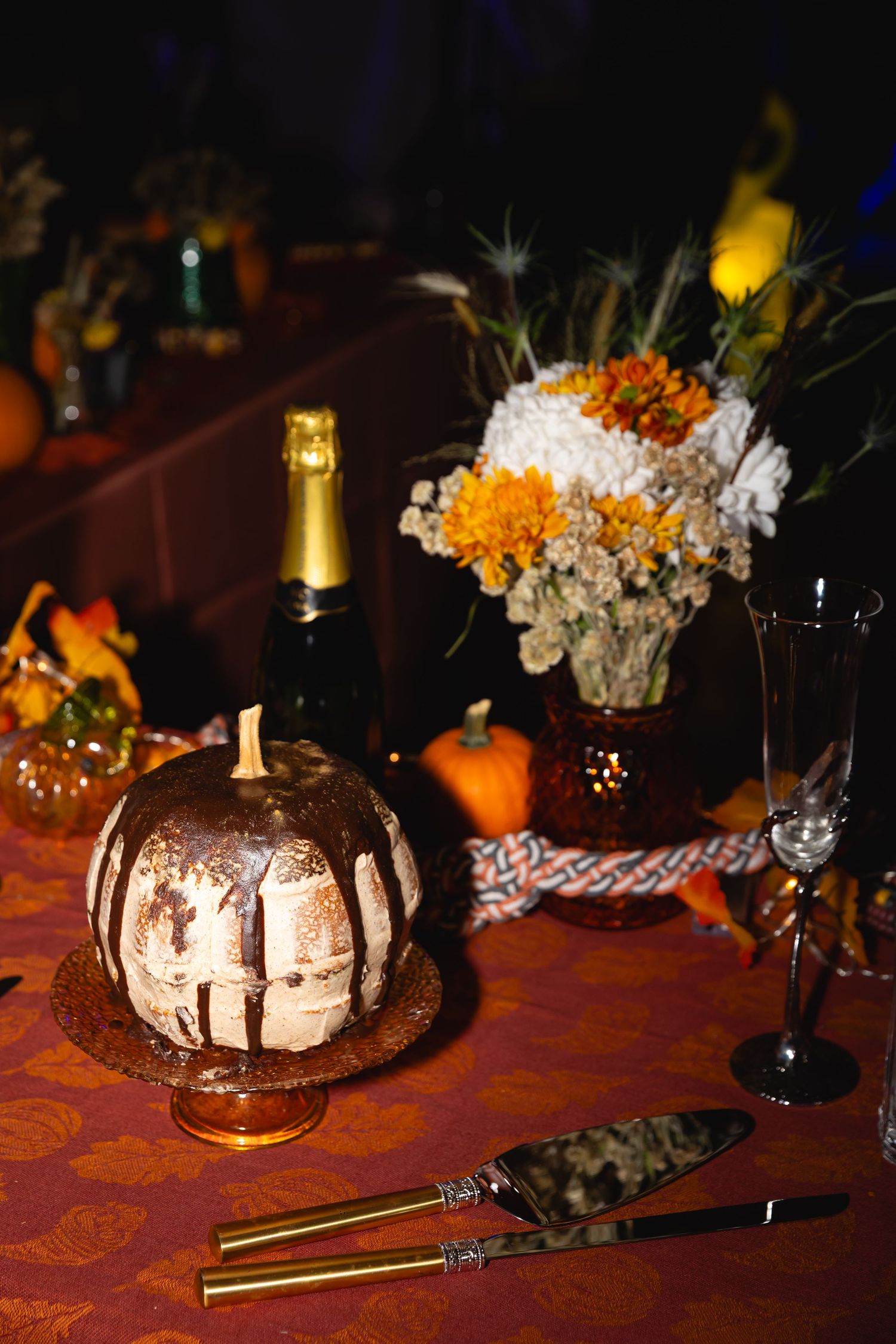 Chocolate drizzled bundt cake decorated with orange and white flowers sits on a dark wooden table with candlelight ambiance.