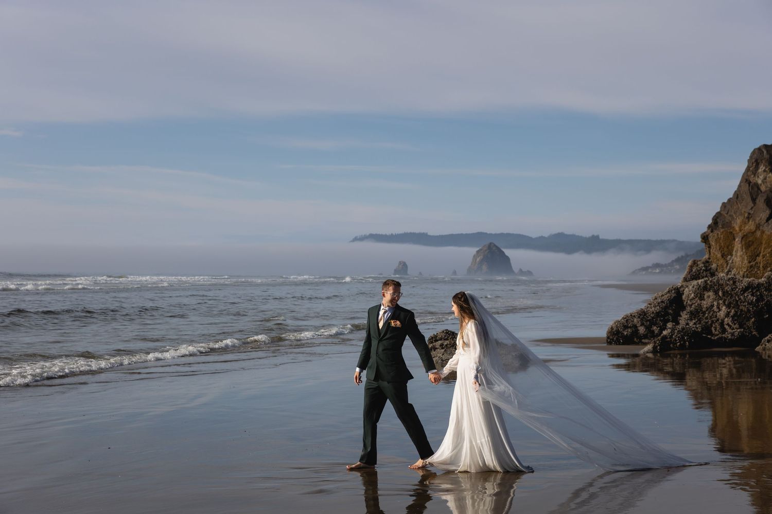 Wedding couple walks along scenic Cannon Beach Oregon with Haystack Rock visible in the misty coastal background.