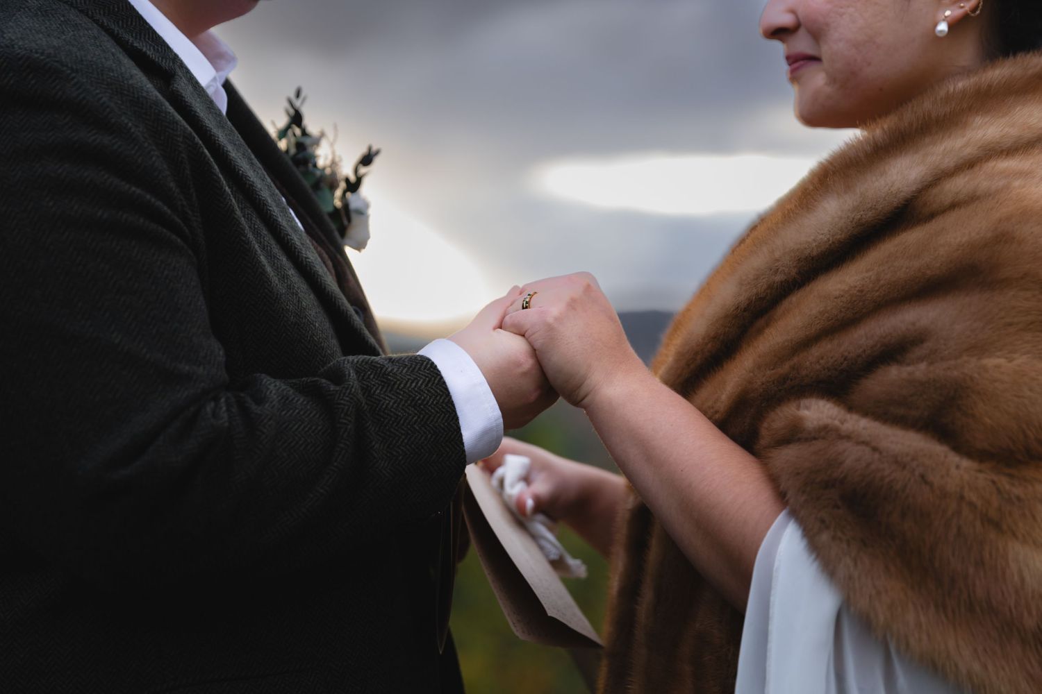 Close-up of wedding couple holding hands with brown fur wrap and suit visible.