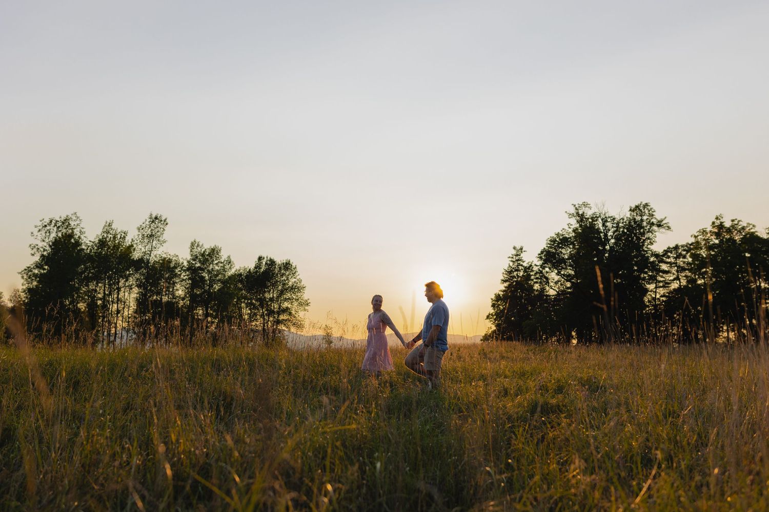 A couple stands together in a sunlit meadow during golden hour with trees silhouetted in the background.