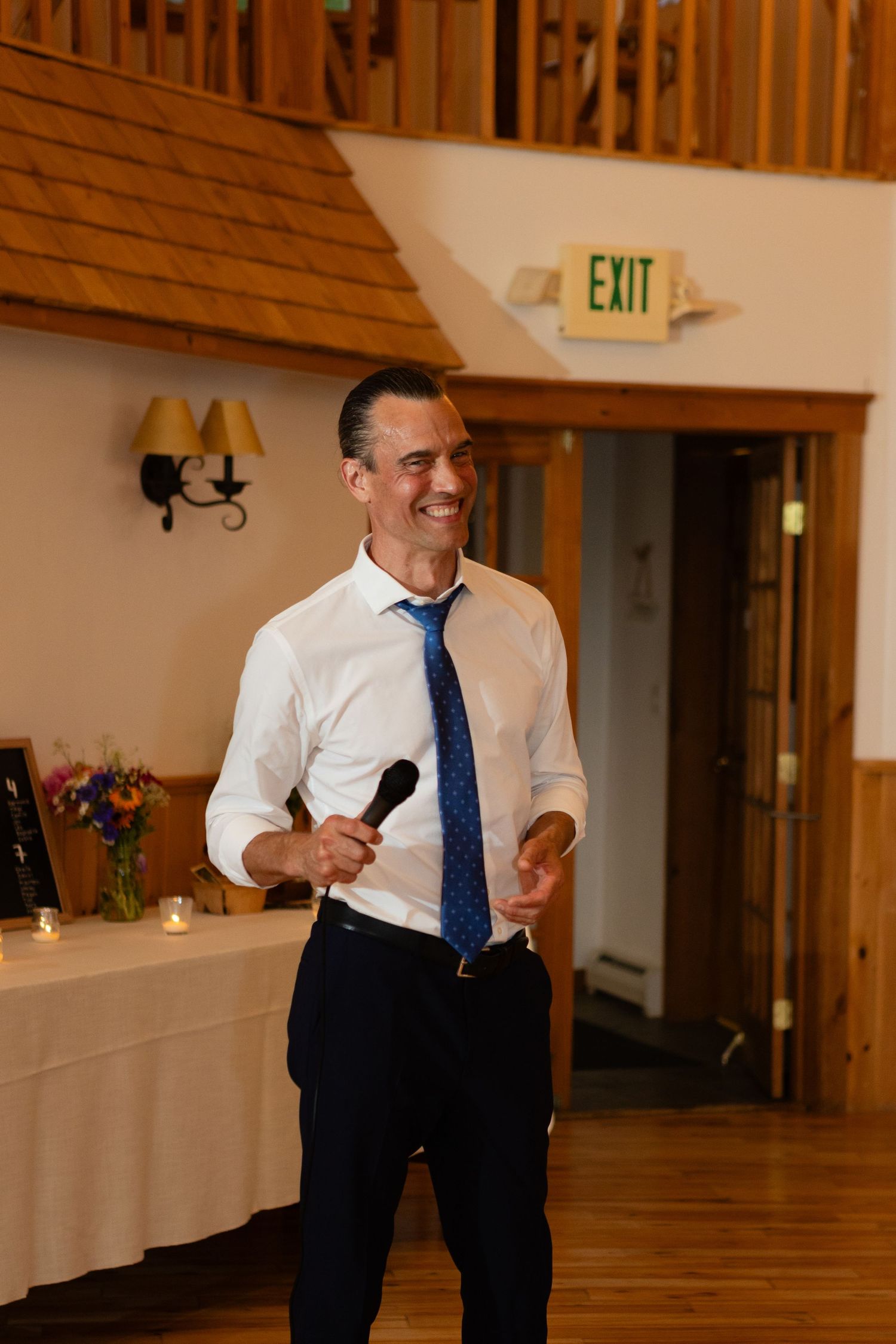 Speaker holding a microphone in formal attire at wooden-trimmed venue.