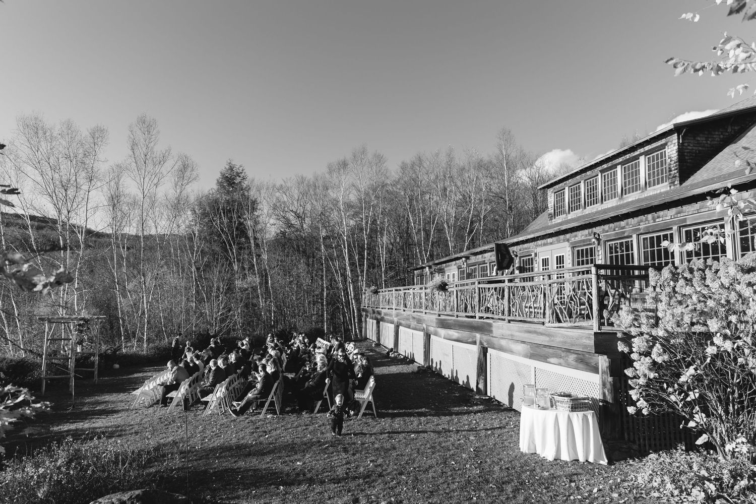 Outdoor wedding venue with guests seated on lawn facing rustic building during fall season.