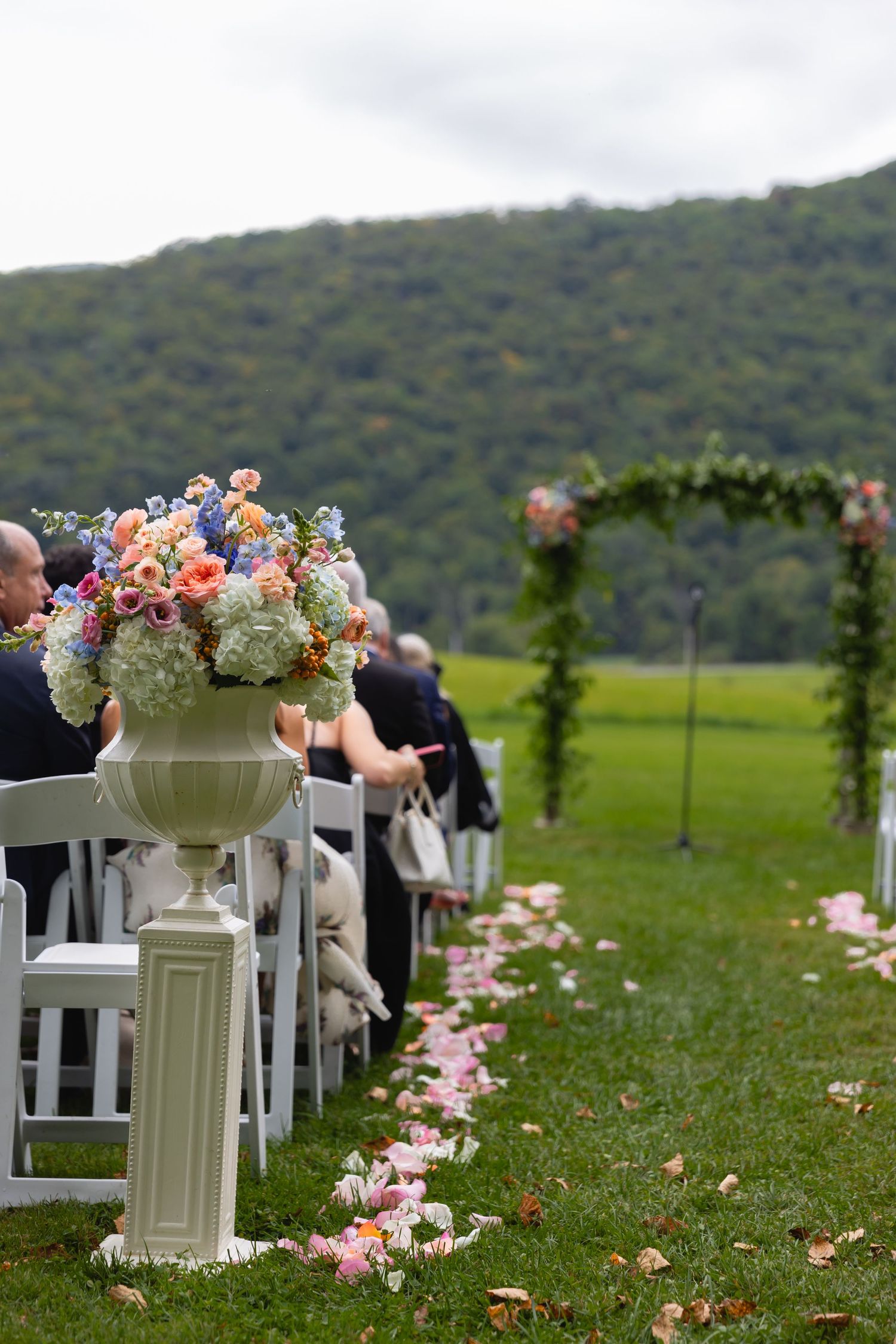 An outdoor wedding ceremony setup with white chairs, floral arrangements, and rose petals along the aisle against a mountain backdrop.