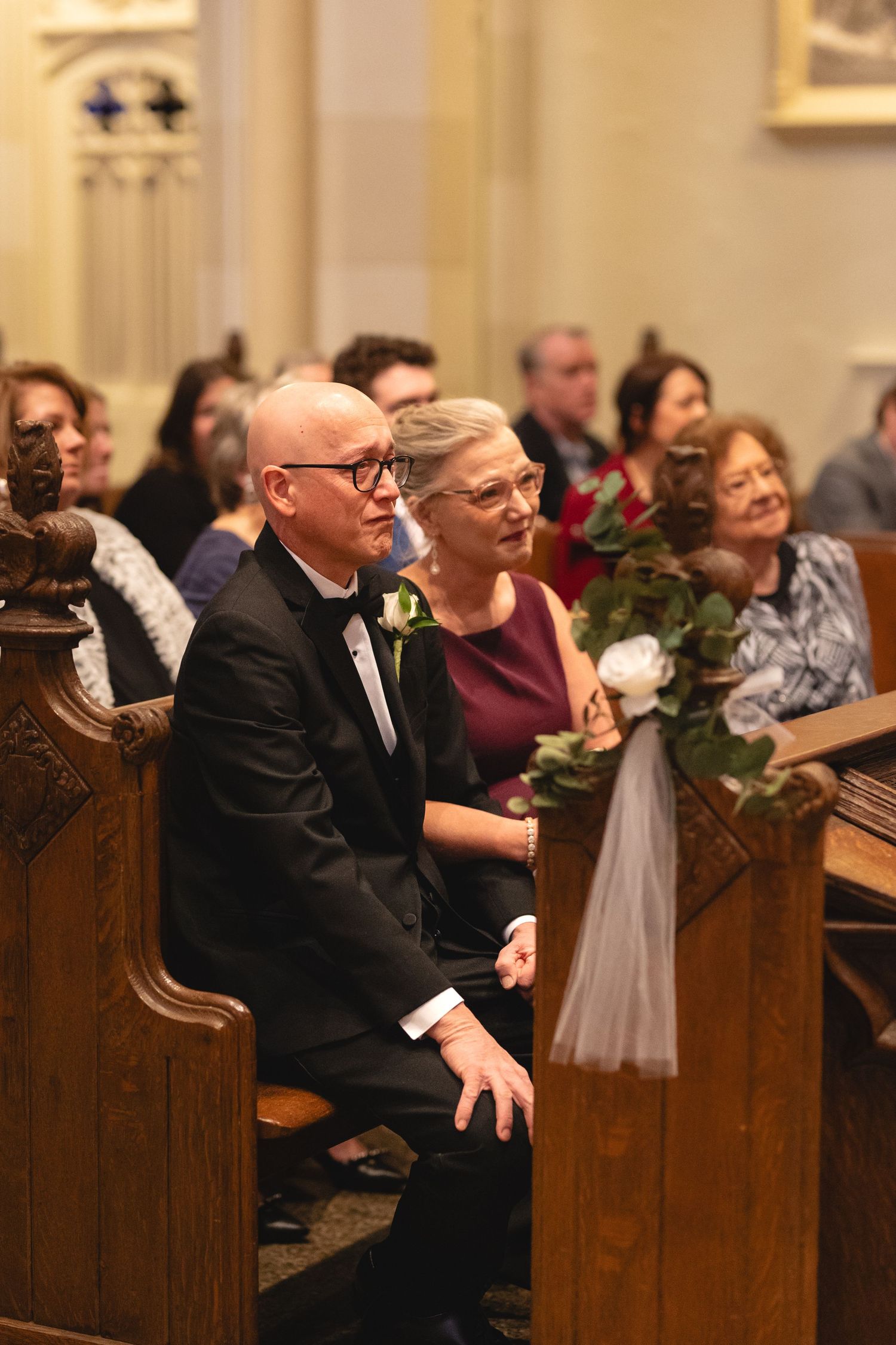 Wedding guests seated in wooden church pews during a ceremony with holiday decorations and white tulle draping.