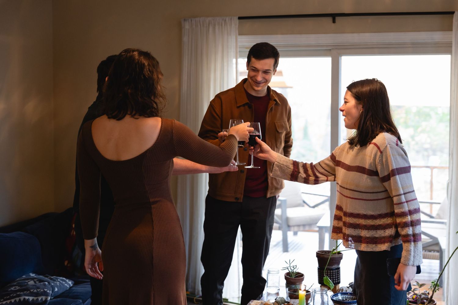 Three friends gather in a living room toasting drinks together beside a window during a casual social gathering.