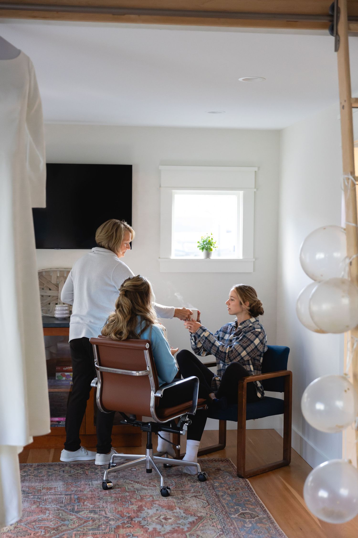 Three people sit and chat in a cozy home office with white walls, a vintage rug, and round globe pendant lights.
