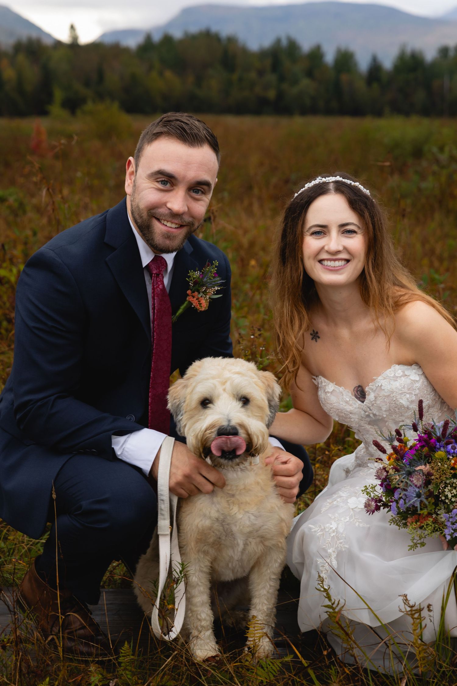 A couple poses with their fluffy white dog amidst mountain scenery while holding a colorful wildflower bouquet.