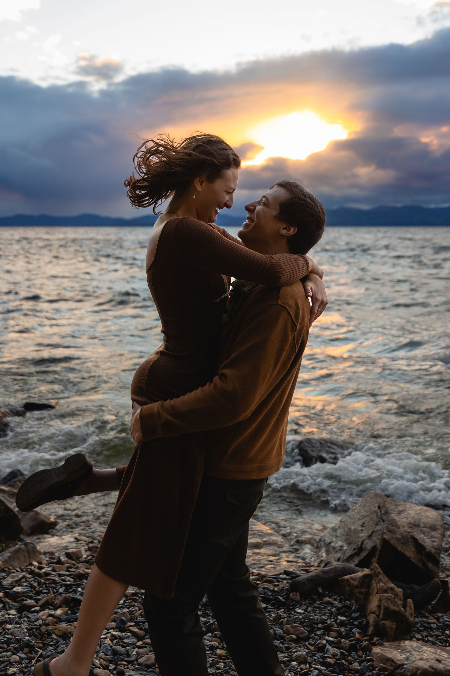A couple embraces on a rocky beach at sunset as dramatic clouds reflect over calm waters in the distance.