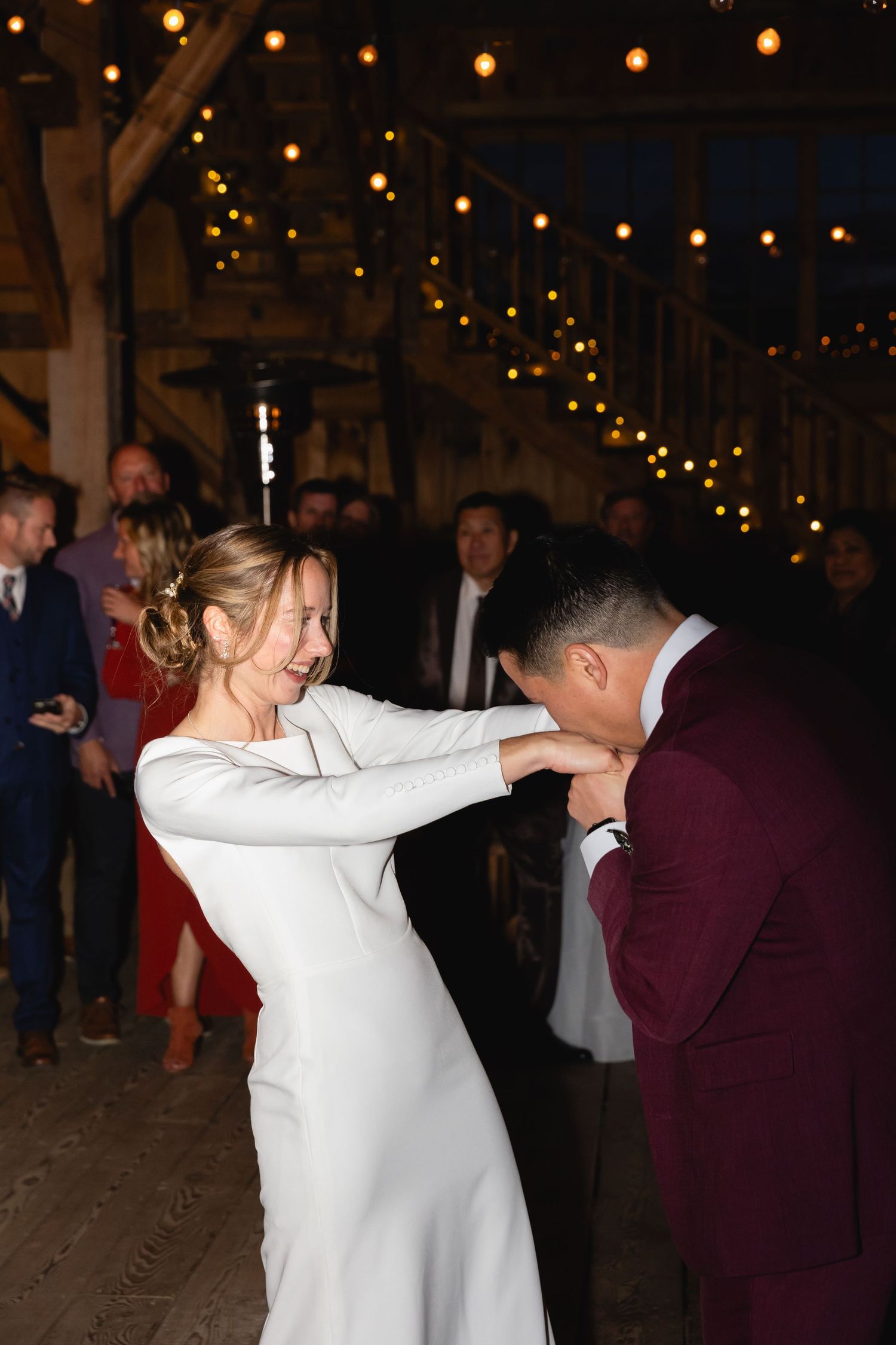 A bride in a sleek white dress and groom in burgundy suit share an intimate moment under string lights at their wedding reception.