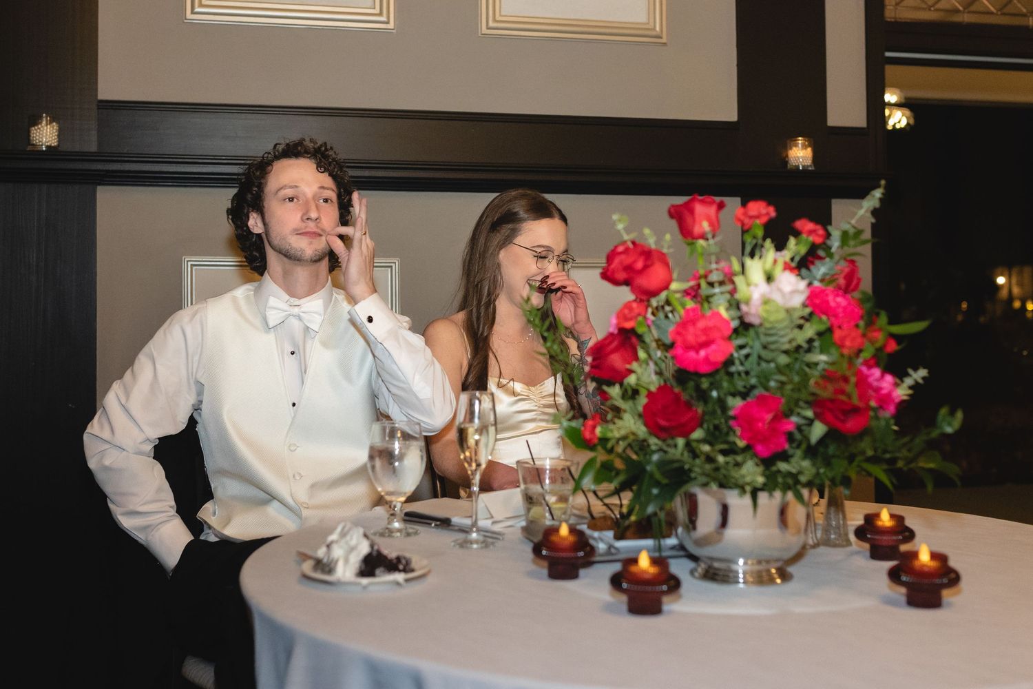 A couple raises their glasses in a toast at a formal dinner table adorned with a beautiful red rose centerpiece.