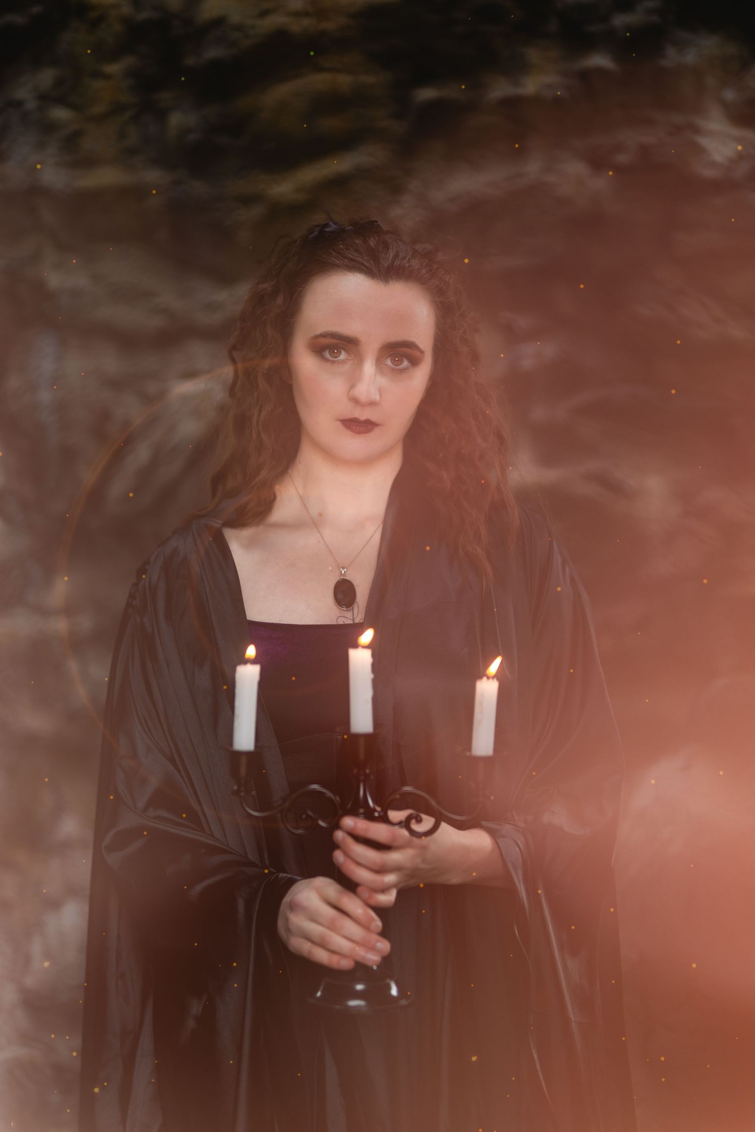 A woman in a black medieval gown holds a candelabra with lit candles in misty light.