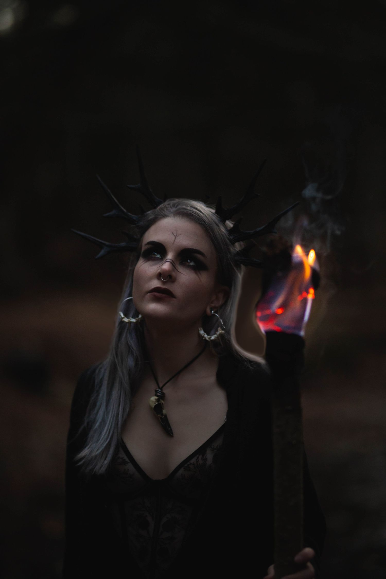 Dark fantasy woman with horned crown holds flaming torch in moody forest setting.