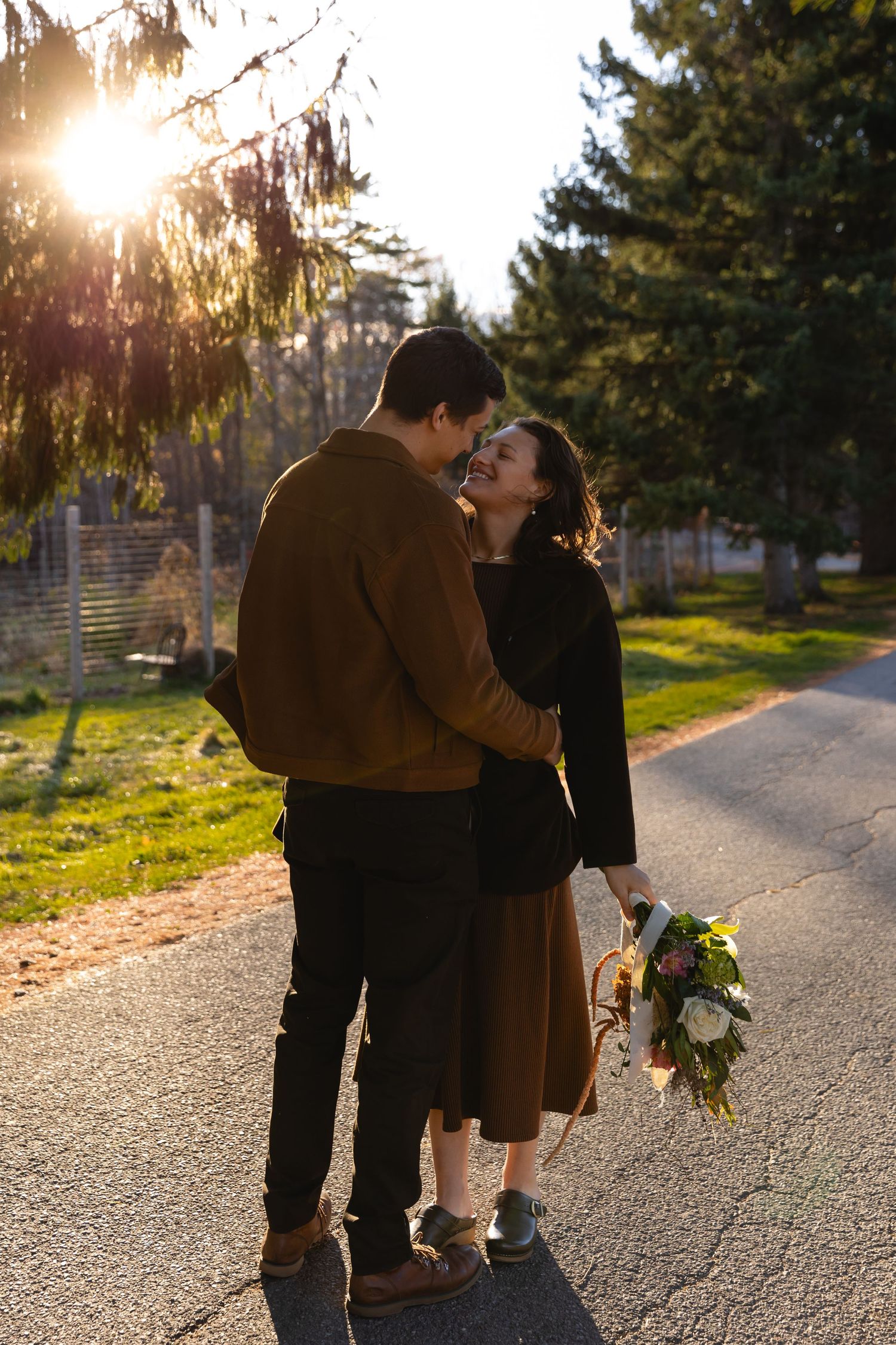 Couple sharing a romantic moment at golden hour on a tree-lined path, woman holding a wildflower bouquet.