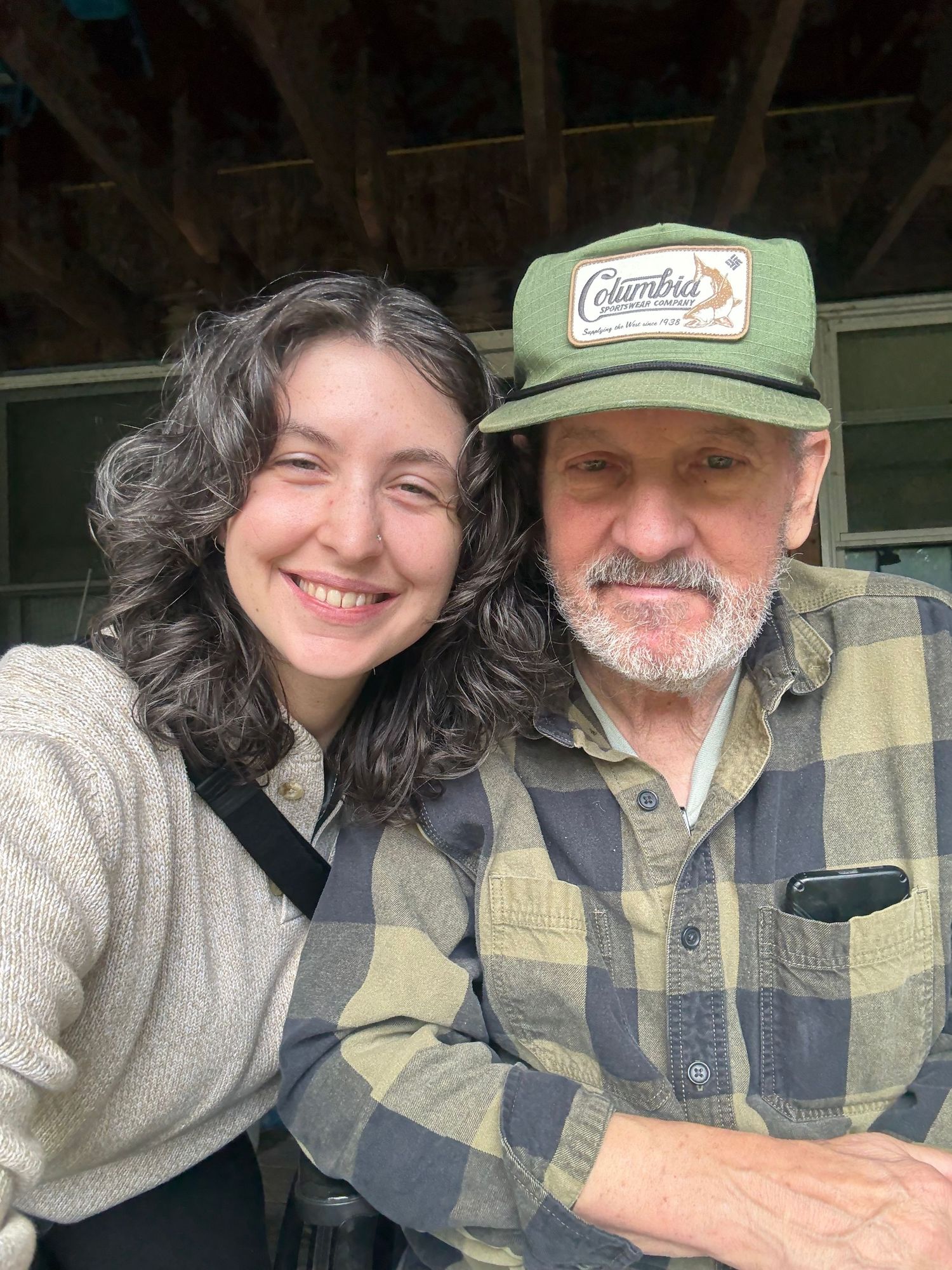 Young woman with curly hair smiling beside an older man in a Columbia cap in a selfie photo.