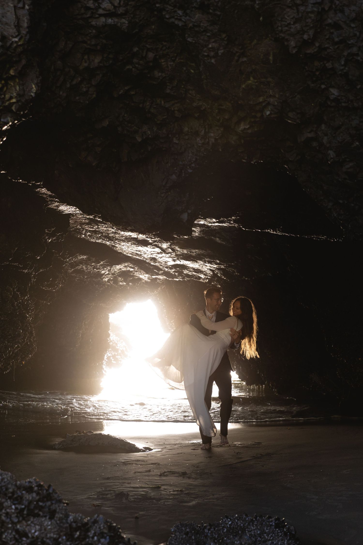 Couple embracing in a dramatic sea cave with glowing light behind them.