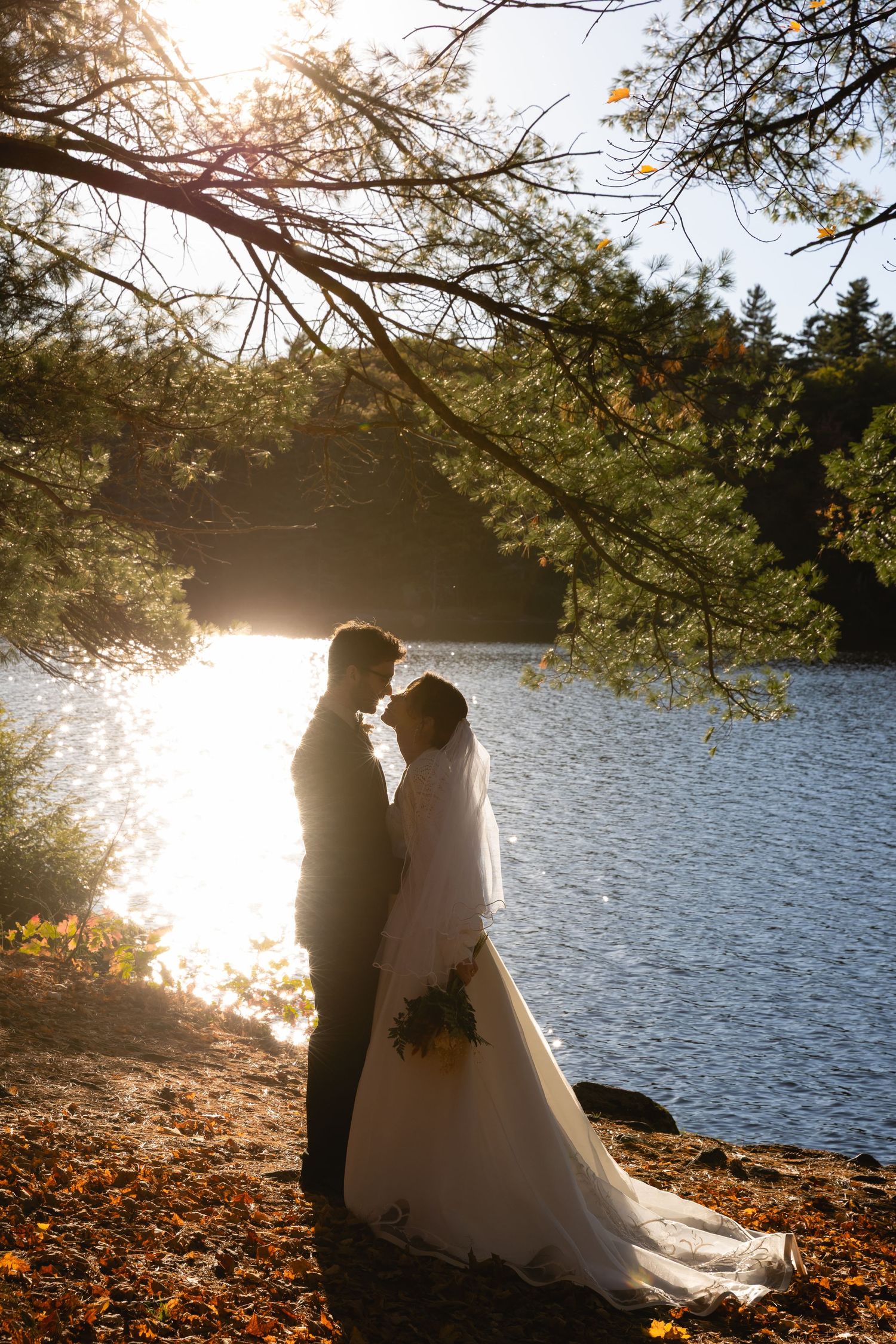 Bride and groom embrace by sunlit lake in golden autumn forest.