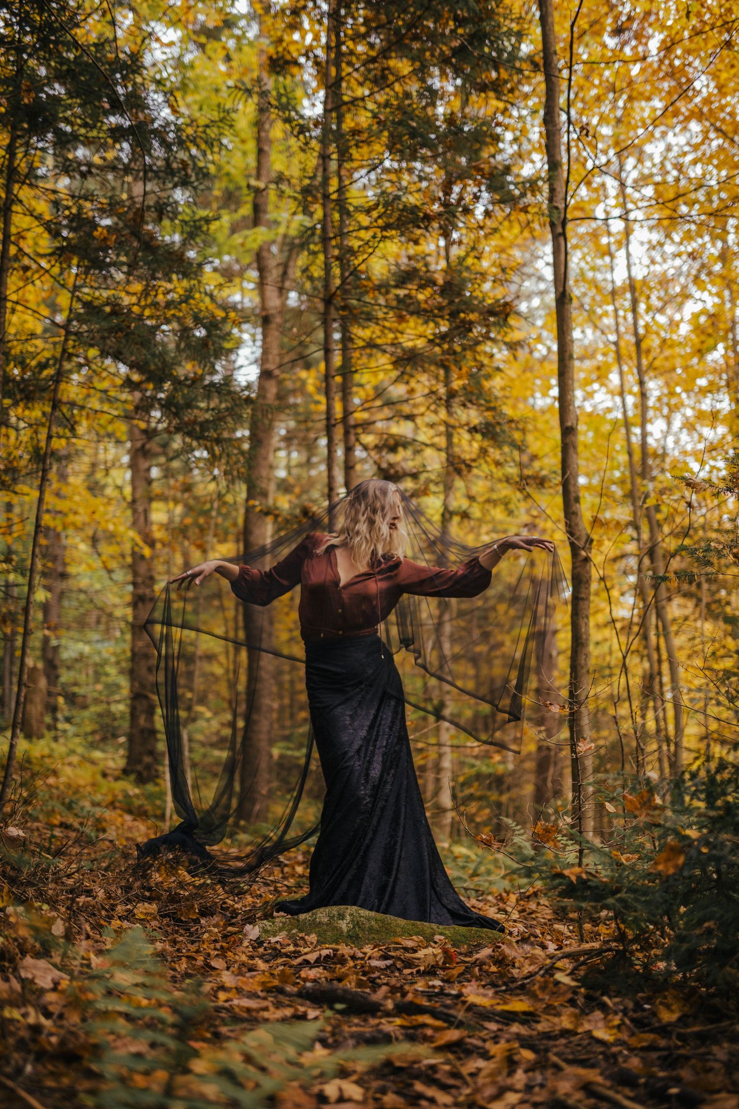 A blonde woman in a black dress dances dramatically in a golden autumn forest holding dark fabric.