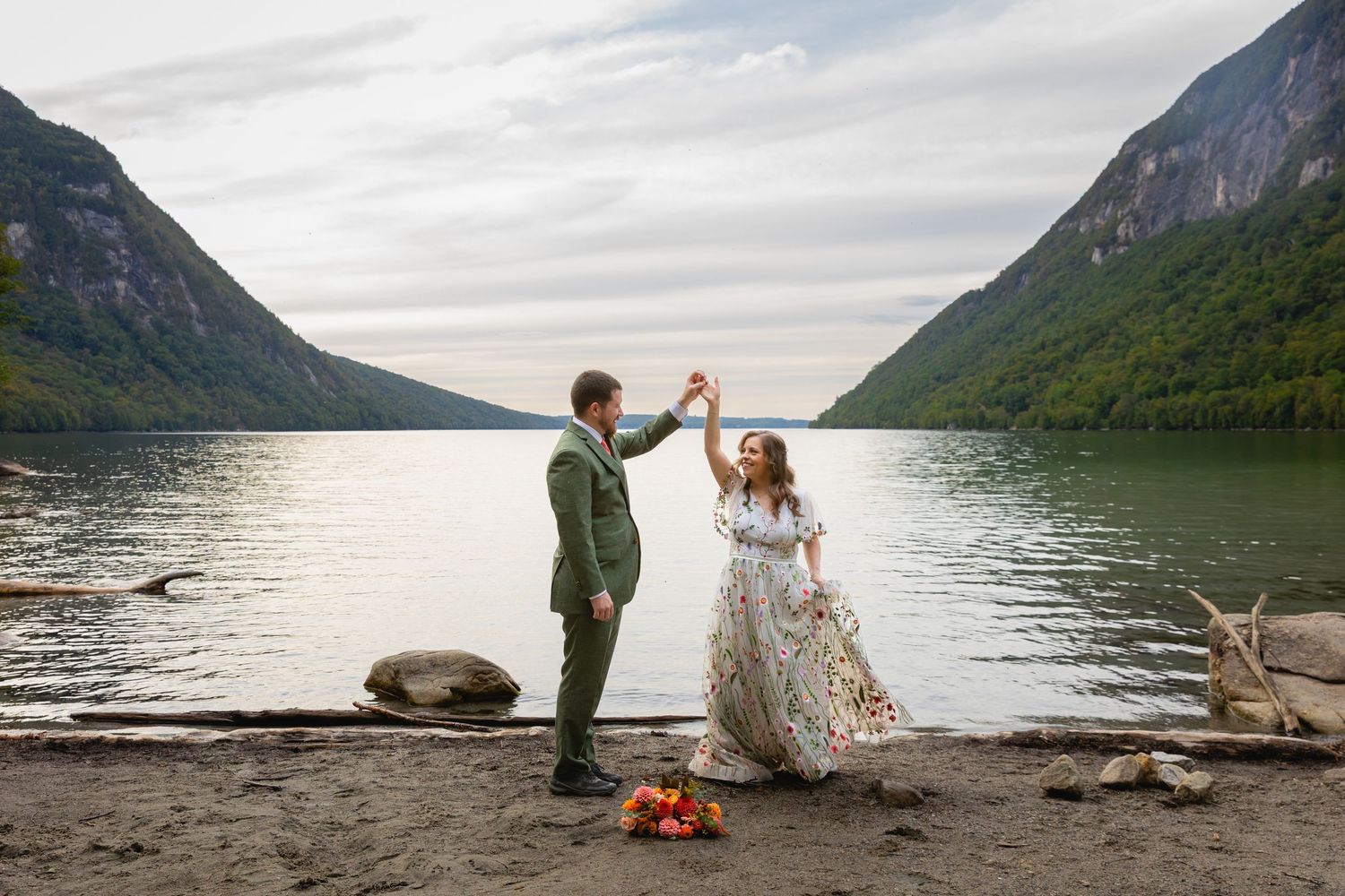 Couple sharing an intimate moment by a serene mountain lake with dramatic cliffs and cloudy skies in the background.