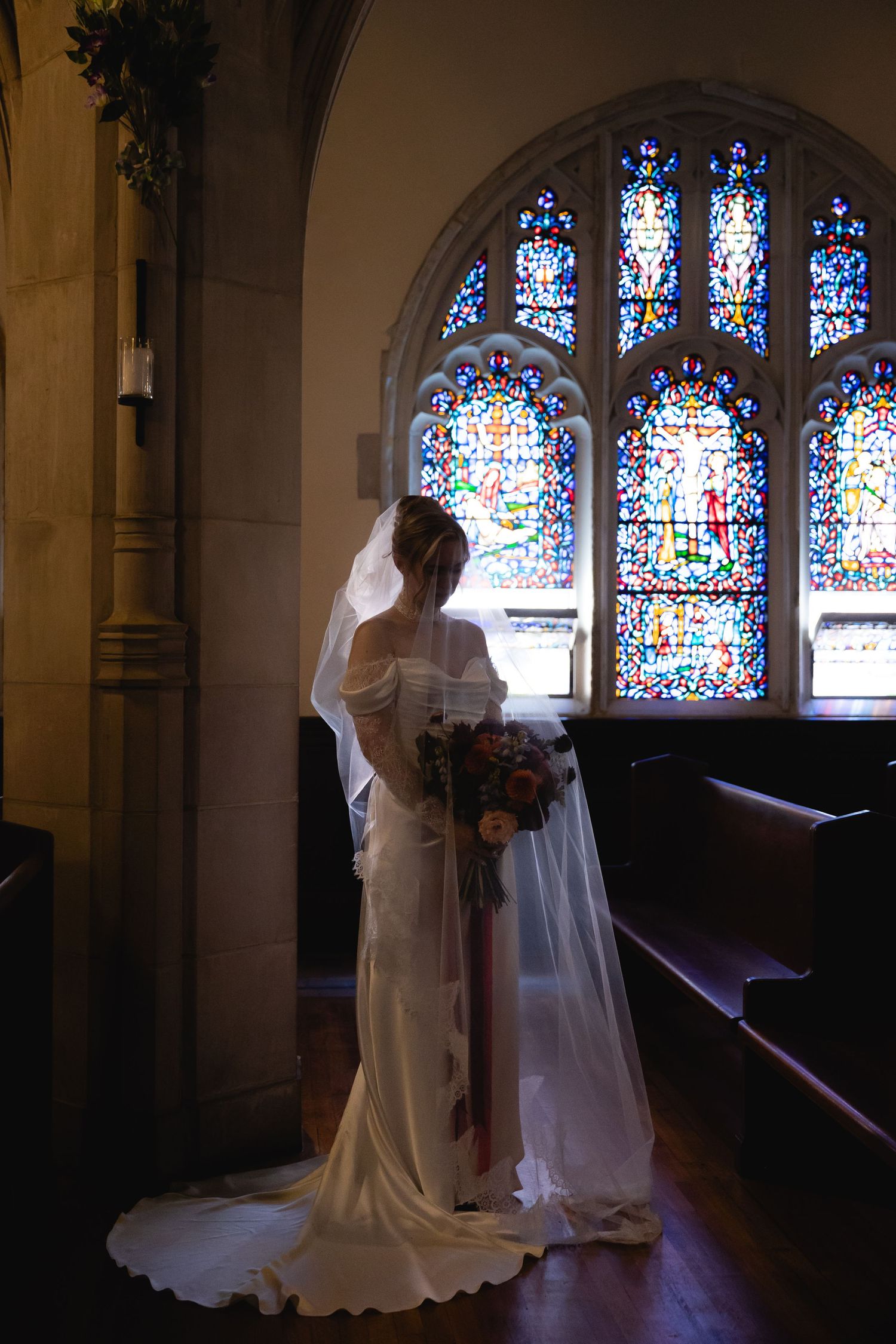 A silhouetted bride stands before a gothic stained glass window in a church, holding a bouquet.