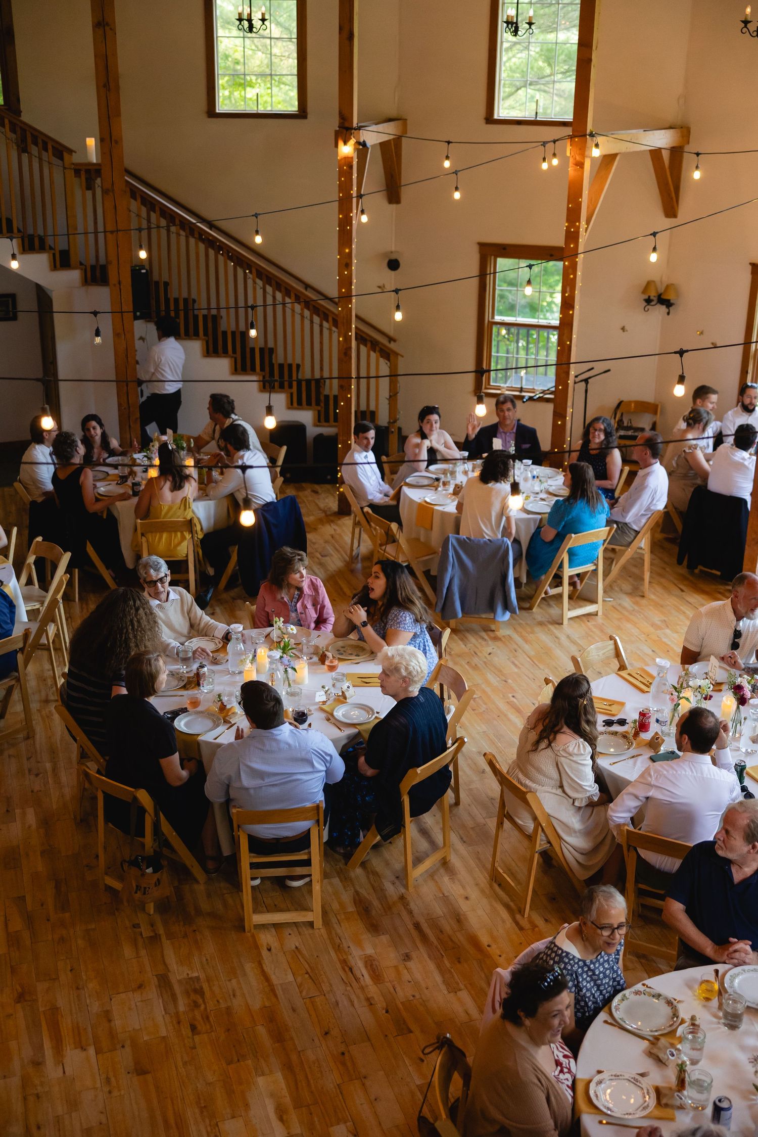 Guests seated at long wooden tables during a rustic indoor reception with string lights overhead and natural light from windows.