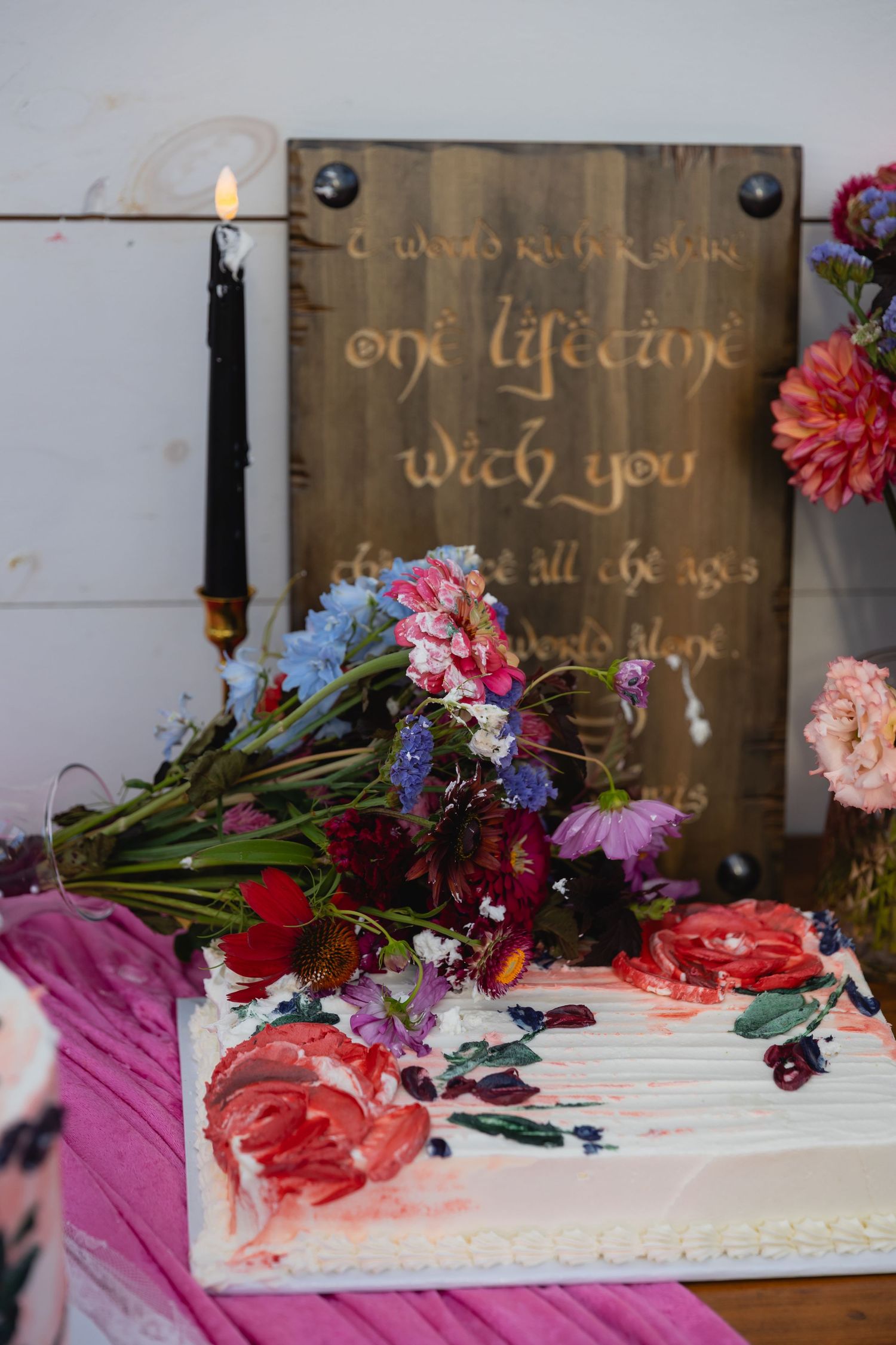 A decorated wedding cake with fresh flowers and a rustic wooden sign display in the background.